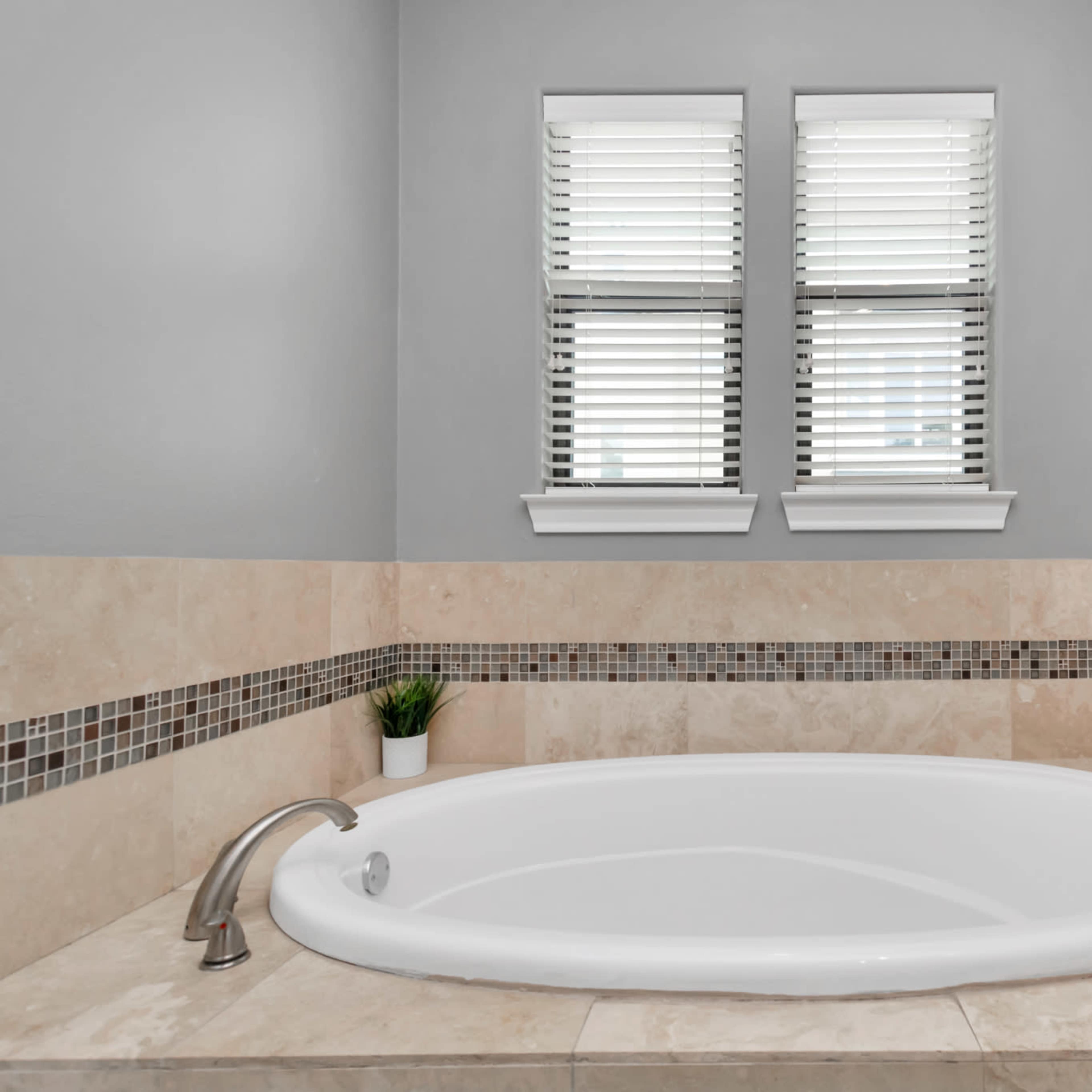 A modern bathroom featuring a white oval bathtub, beige tiled walls, and two windows with blinds.
