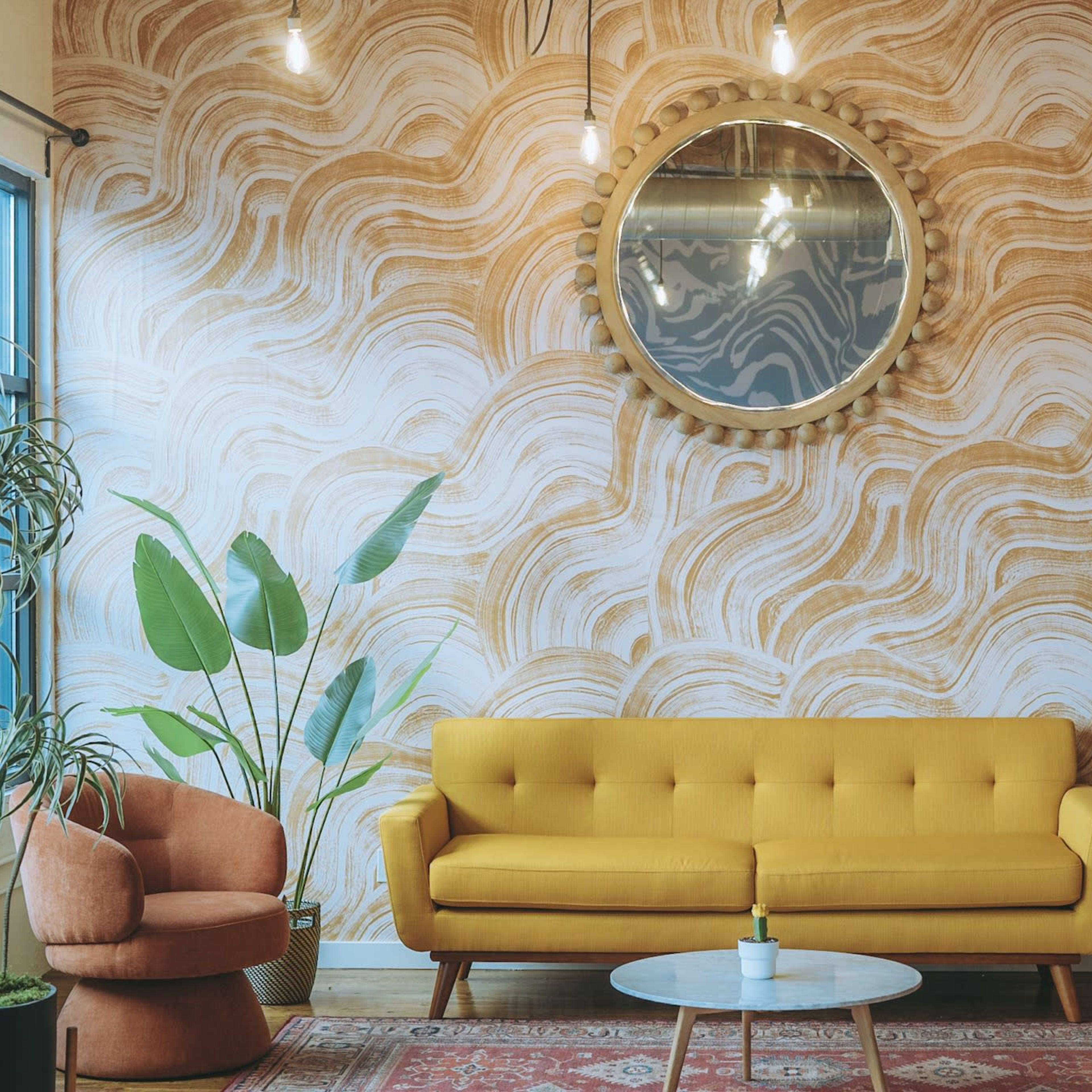 A modern living room featuring a yellow sofa, an orange armchair, a circular mirror, and plants against a wavy patterned wall.