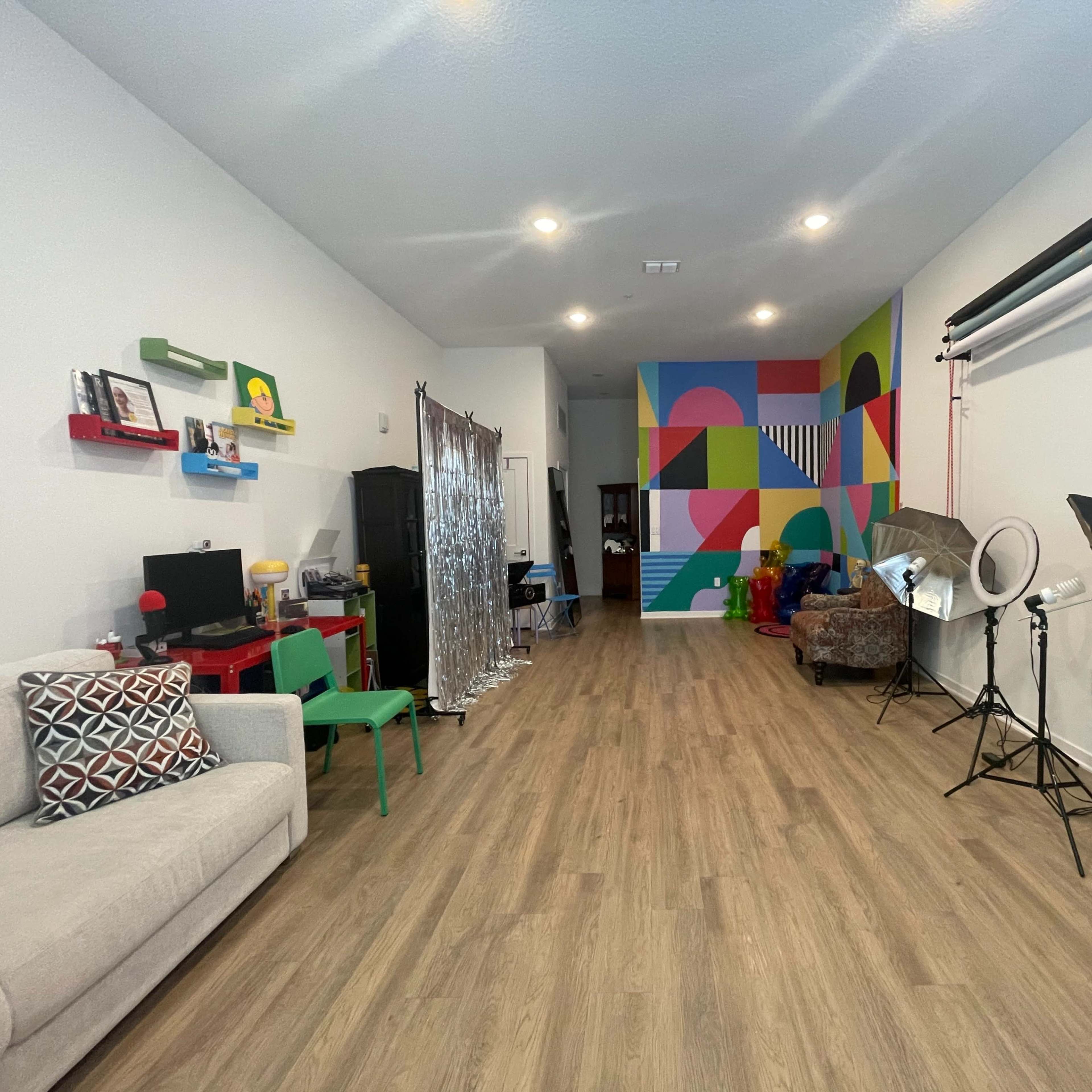 A spacious room with a light wood floor, featuring a colorful mural on one wall, furniture including a couch and chairs, and various equipment for photography.