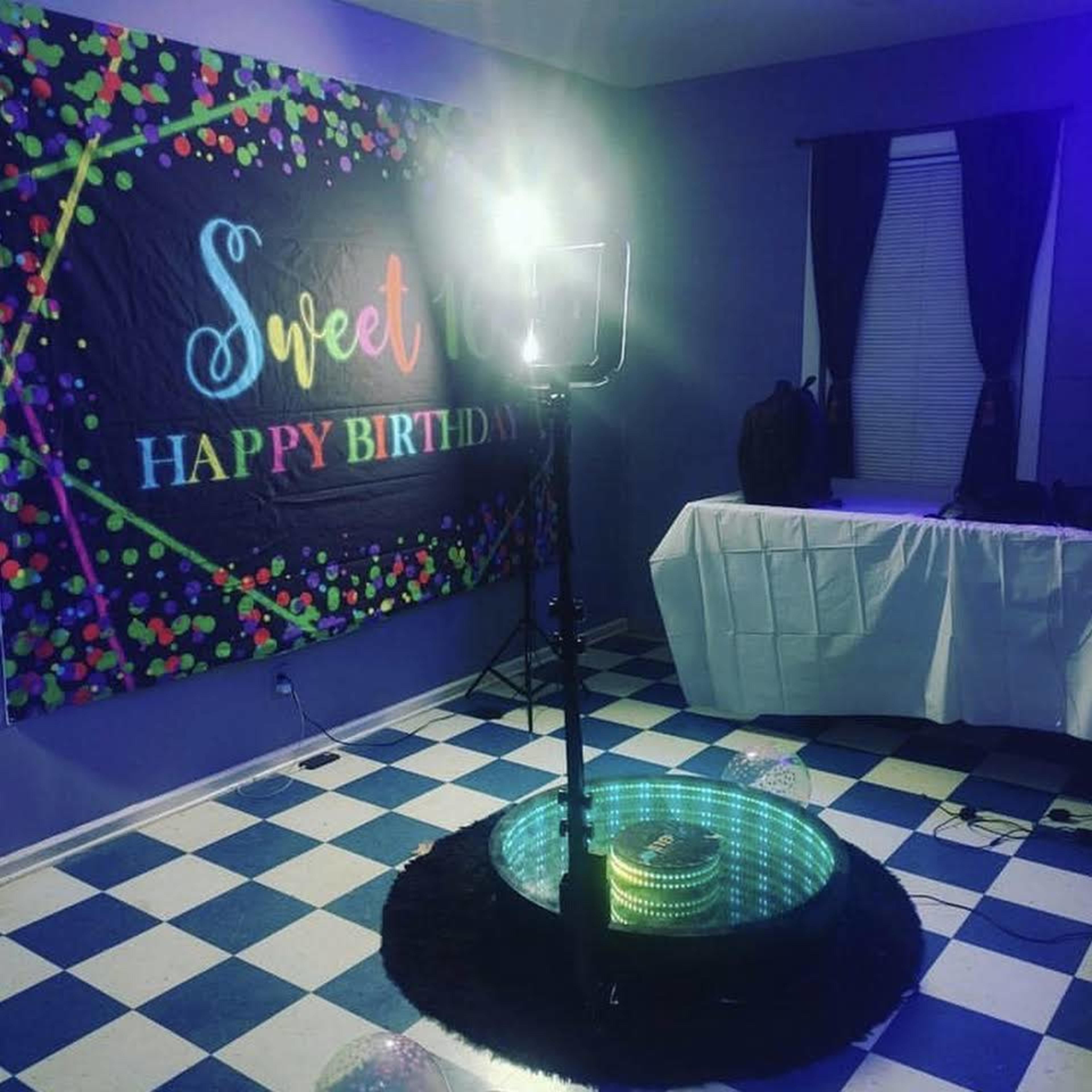 A birthday celebration setup in a room featuring a colorful backdrop, a light stand, and a decorated table with a checkered floor.
