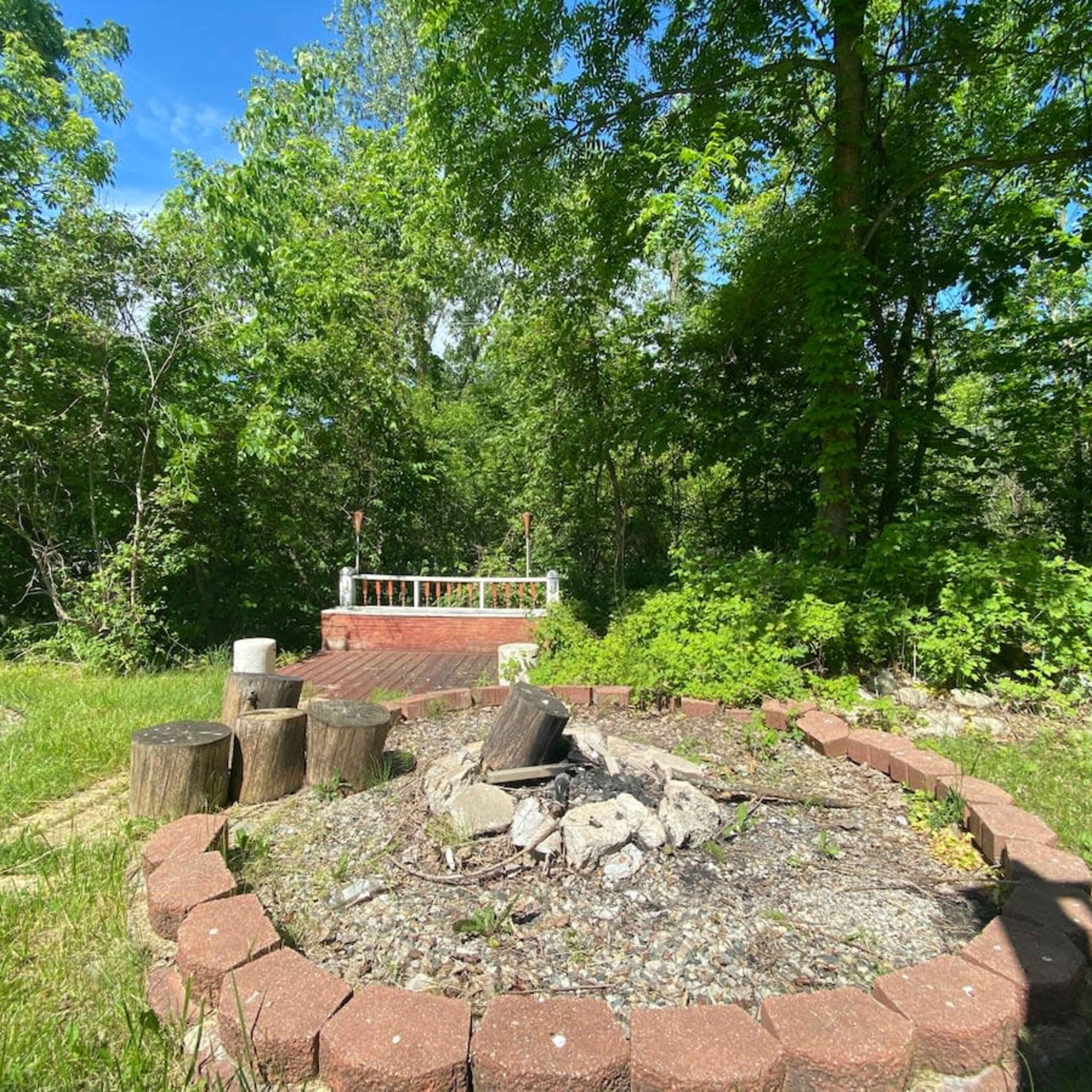 A stone fire pit surrounded by wooden stumps is situated in a grassy area, with trees and greenery in the background.