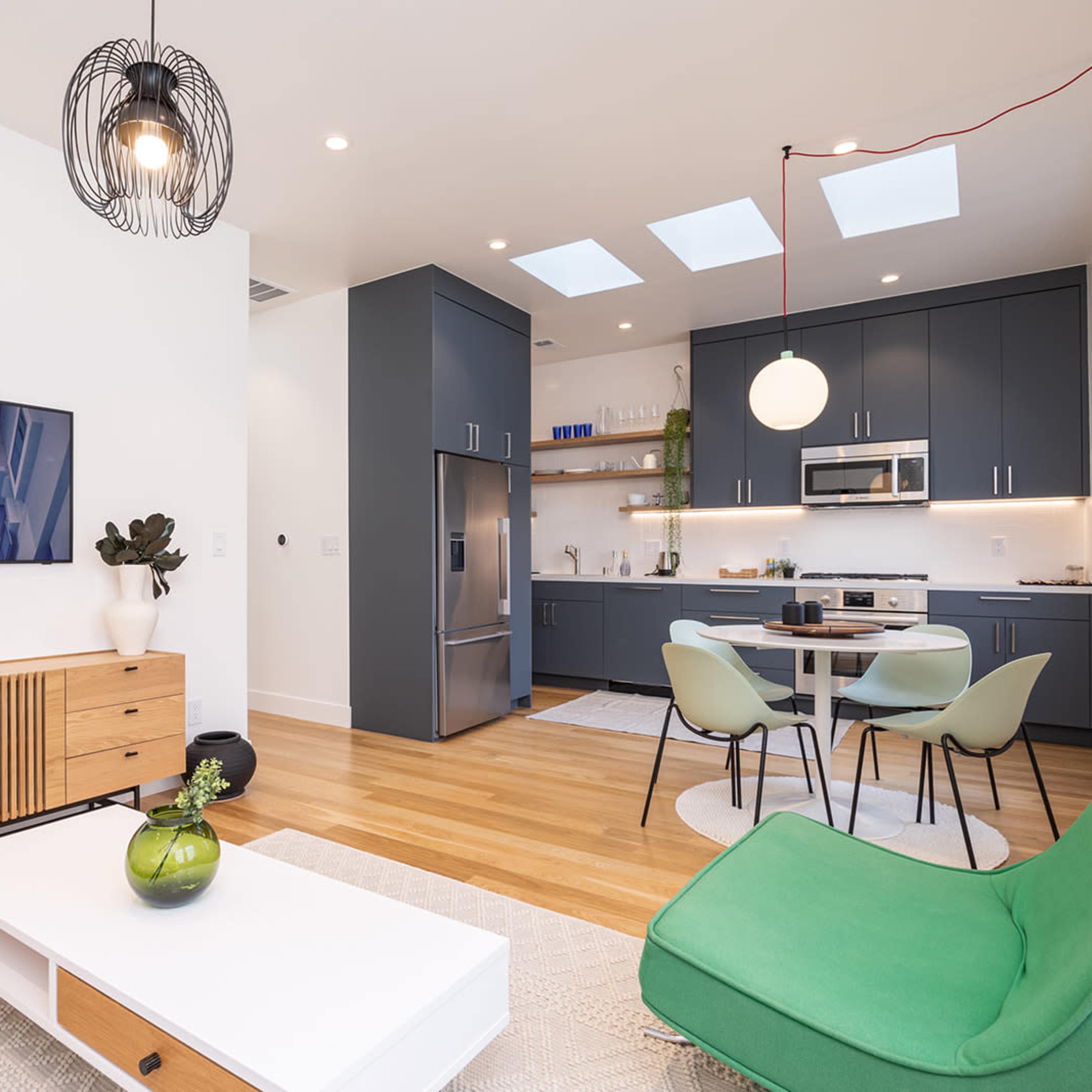A modern living space featuring a green sofa, a wooden media console, a kitchen area with dark cabinets, and a dining table with chairs under skylights.