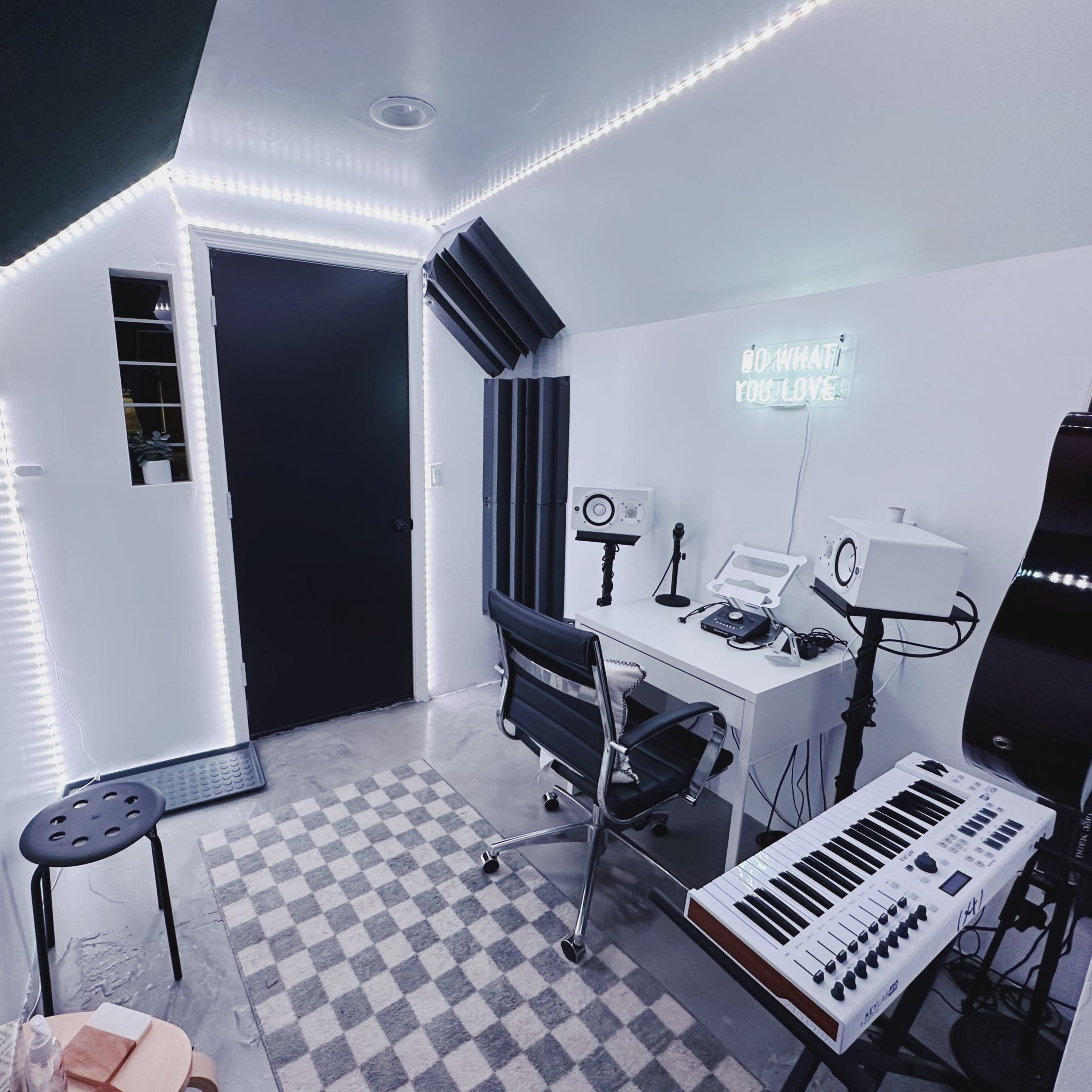 A small music studio featuring a desk with equipment, a keyboard, and acoustic panels, all illuminated by LED lights along the walls.