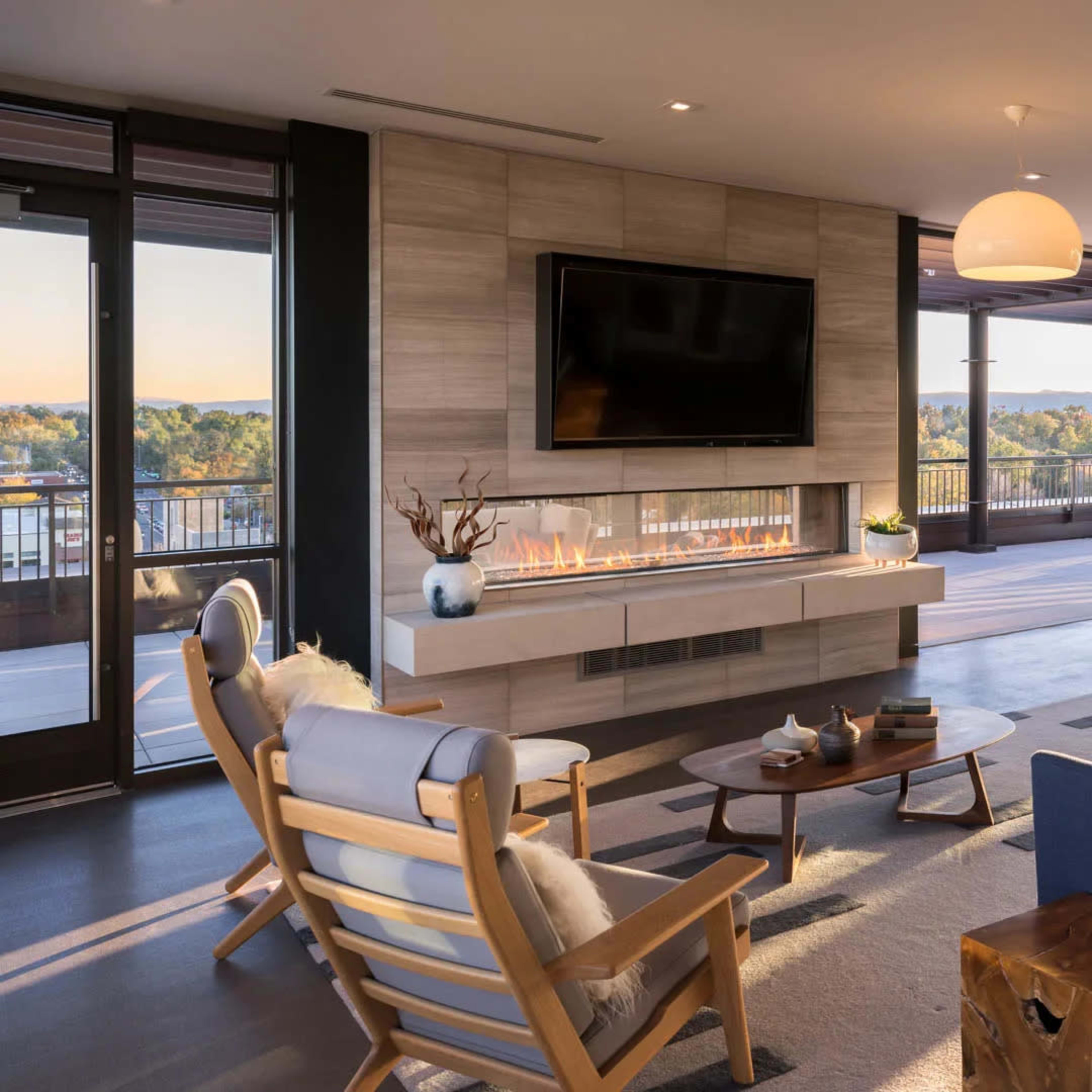 A modern living room with large windows, a television mounted on the wall, a fireplace, and contemporary furniture, including a wooden coffee table and lounge chairs.