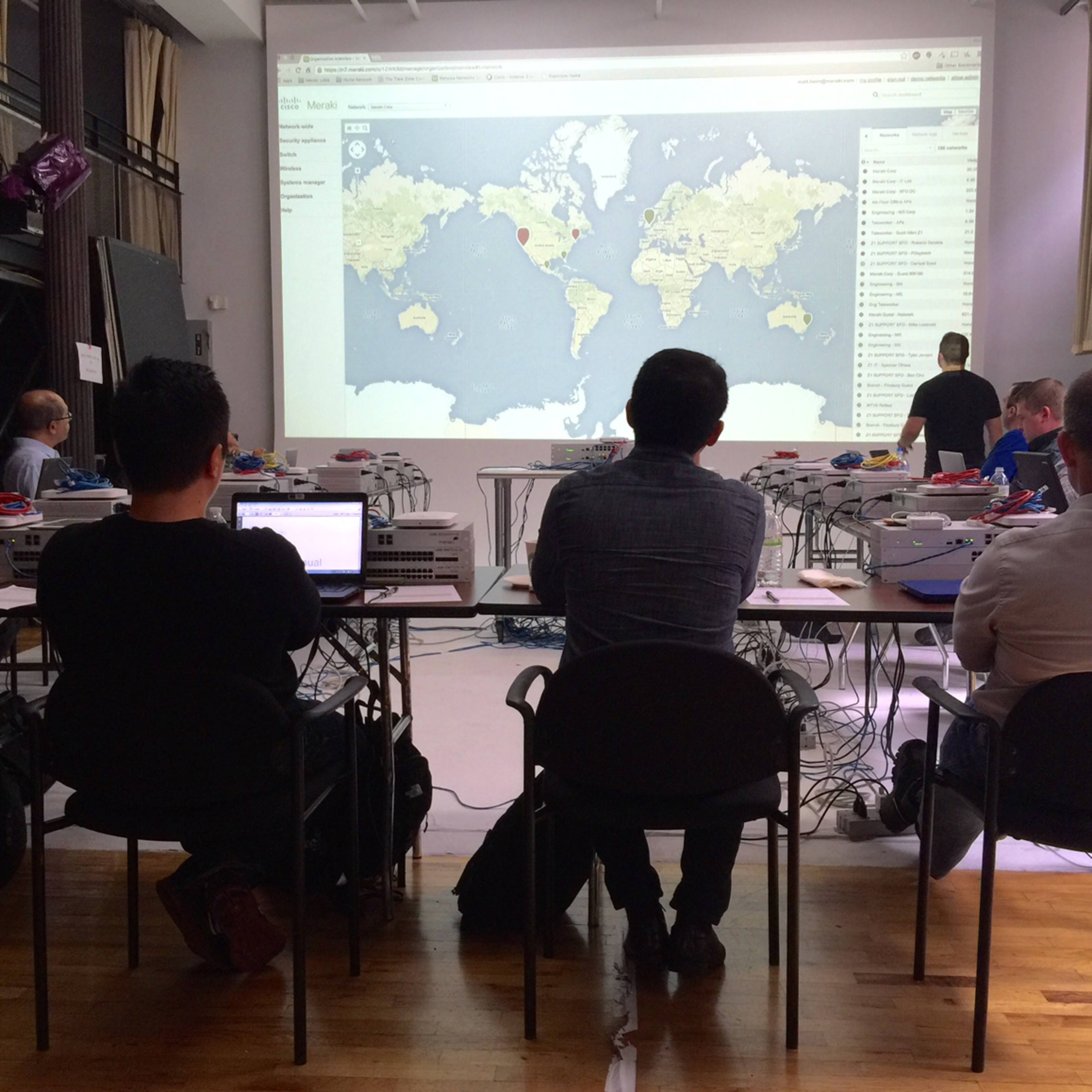 A group of people sits at a long table facing a projected world map on a screen in a classroom setting.