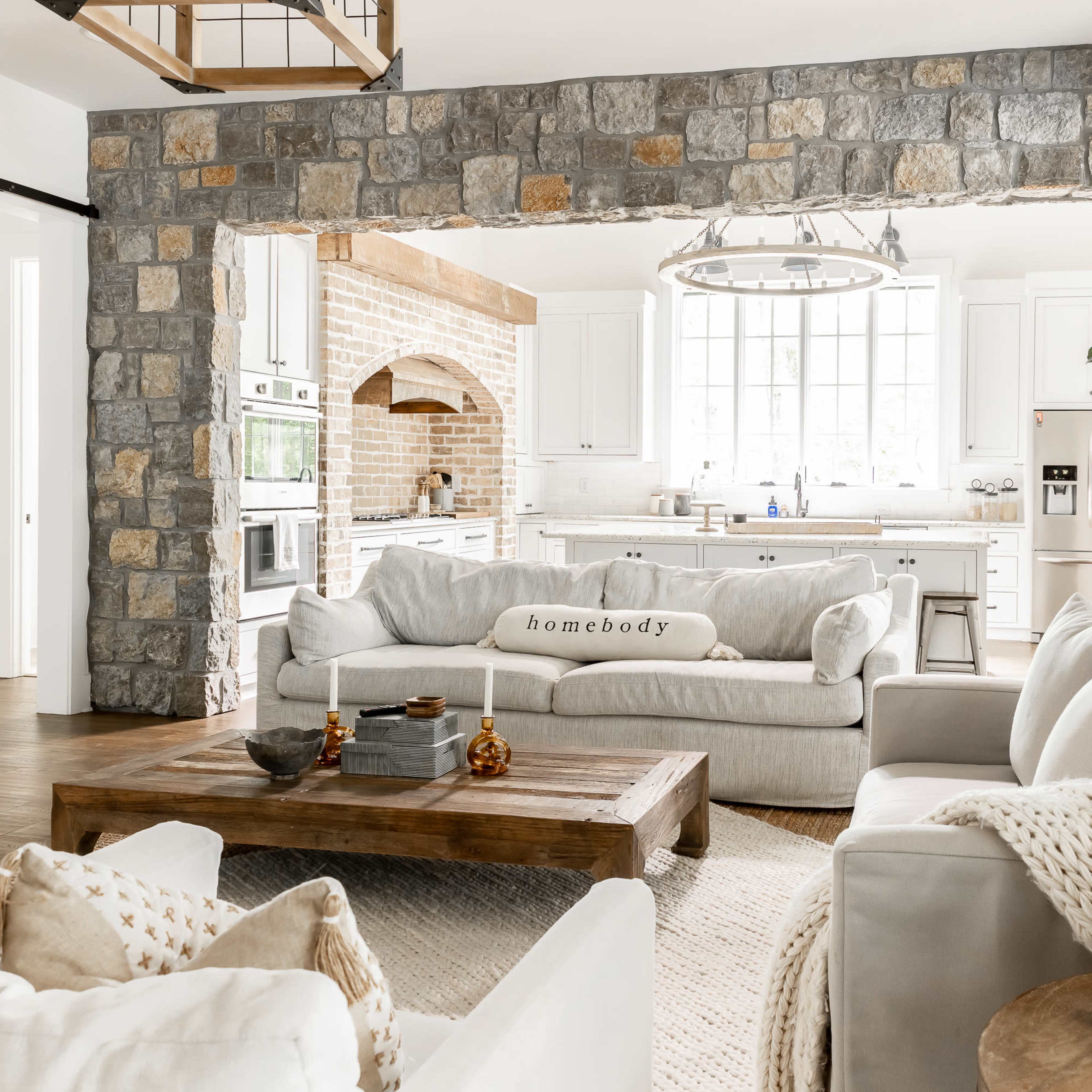 A modern kitchen and living area with a stone archway, featuring a soft gray sofa, a wooden coffee table, and a bright, open layout with large windows.