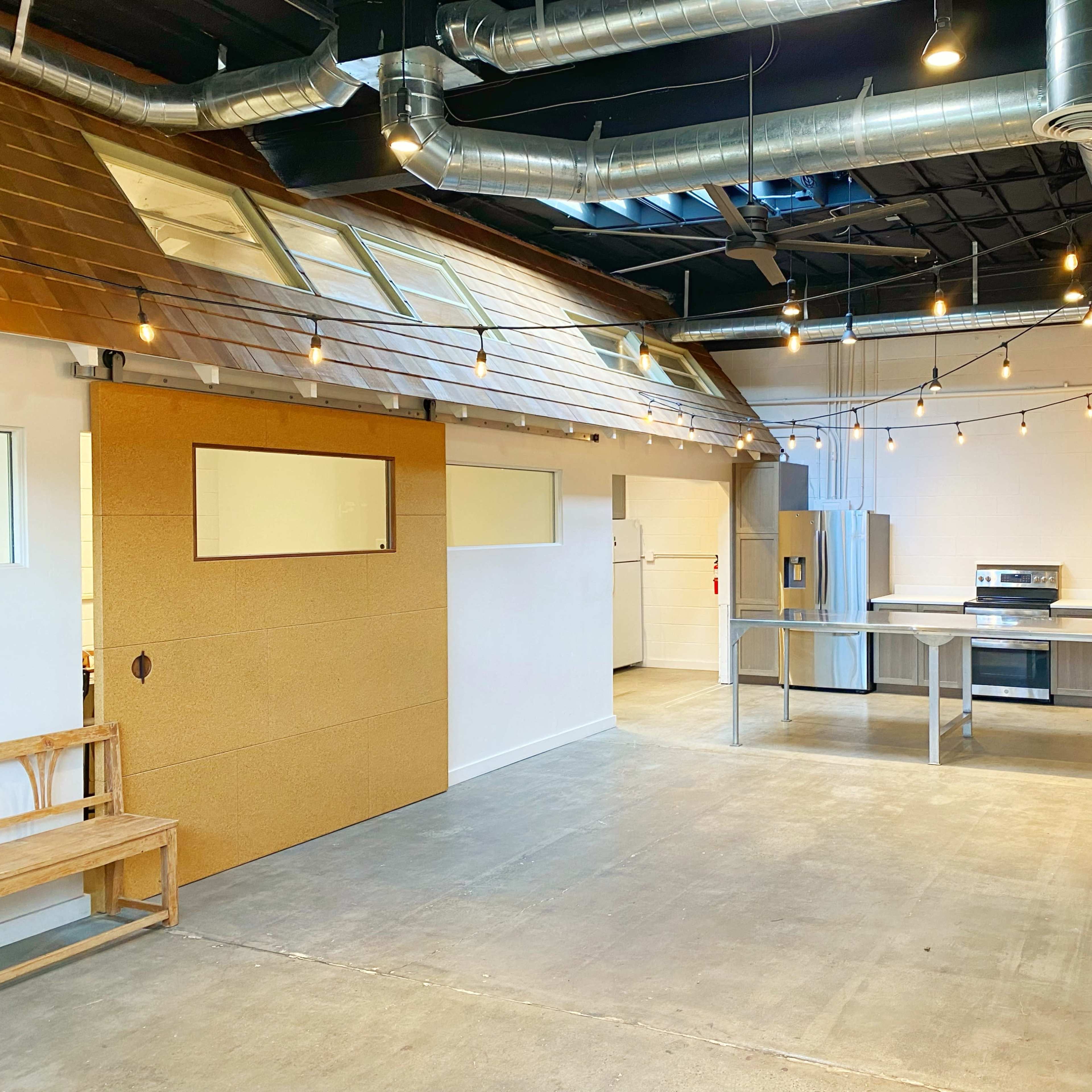 A spacious, modern kitchen area with stainless steel appliances, a wooden bench, and overhead string lights in an industrial-style room.