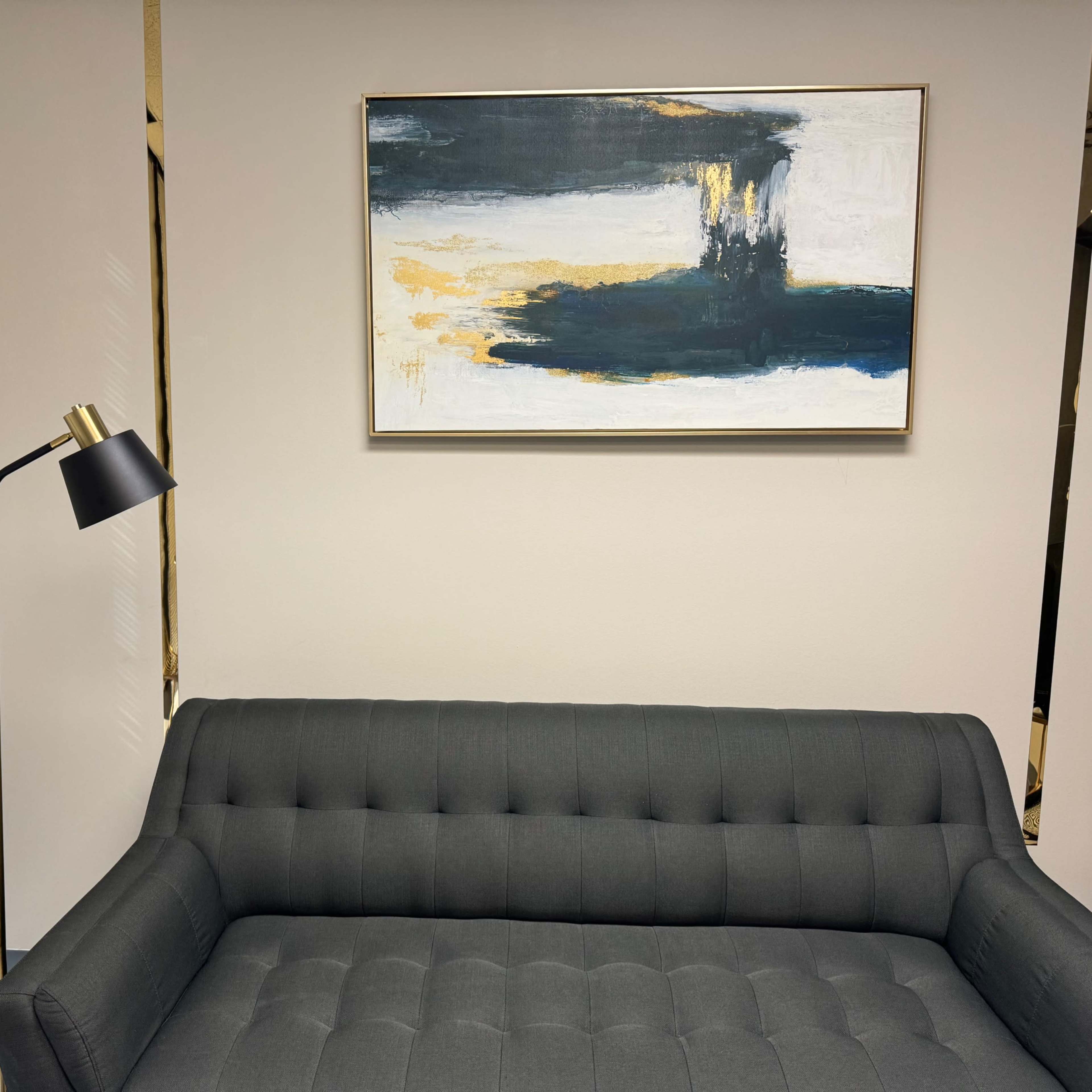 A modern office interior featuring a gray tufted sofa, a floor lamp, and an abstract painting hung on the wall.