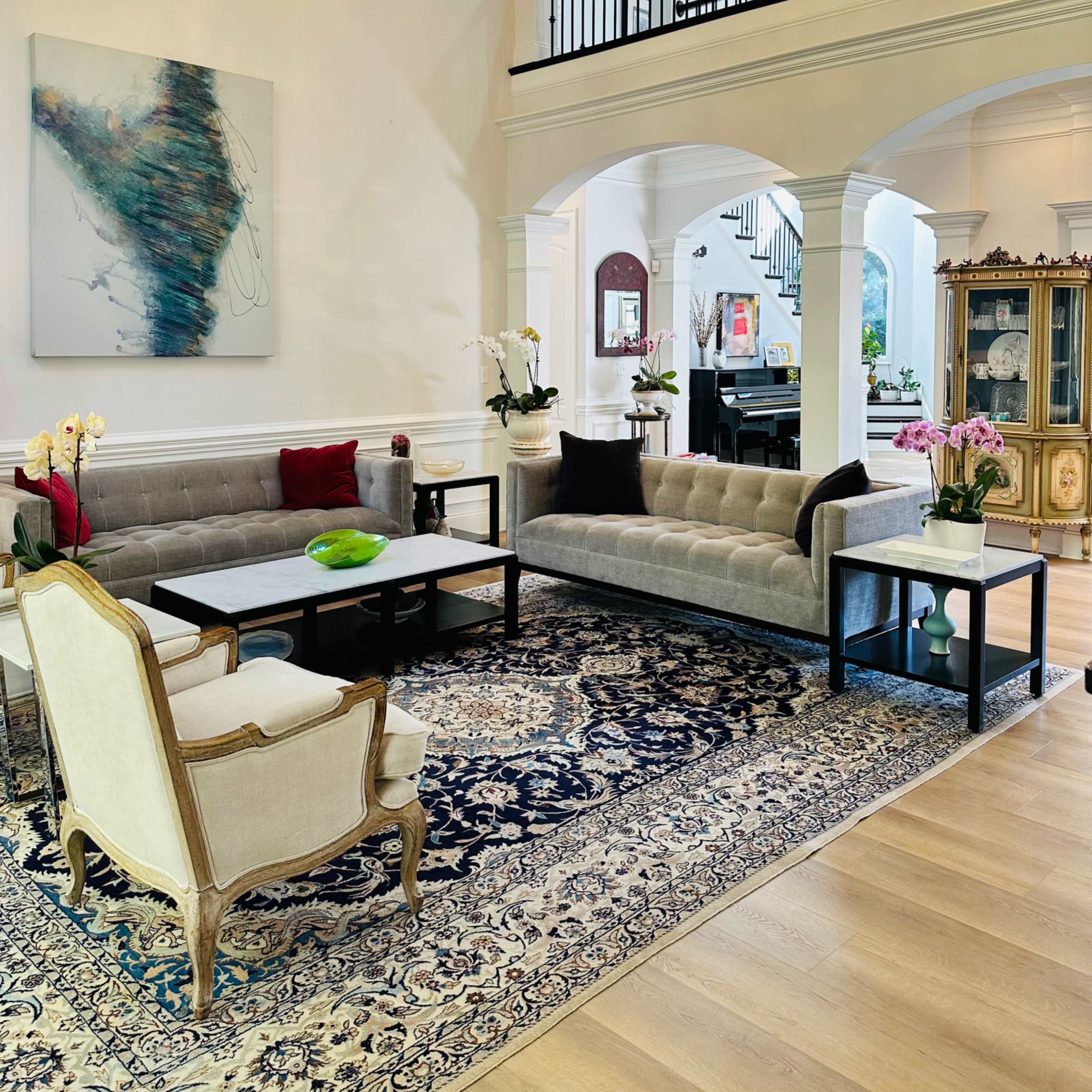A spacious living room featuring a chandelier, a large artwork on the wall, two gray sofas, a marble coffee table, and a detailed rug.