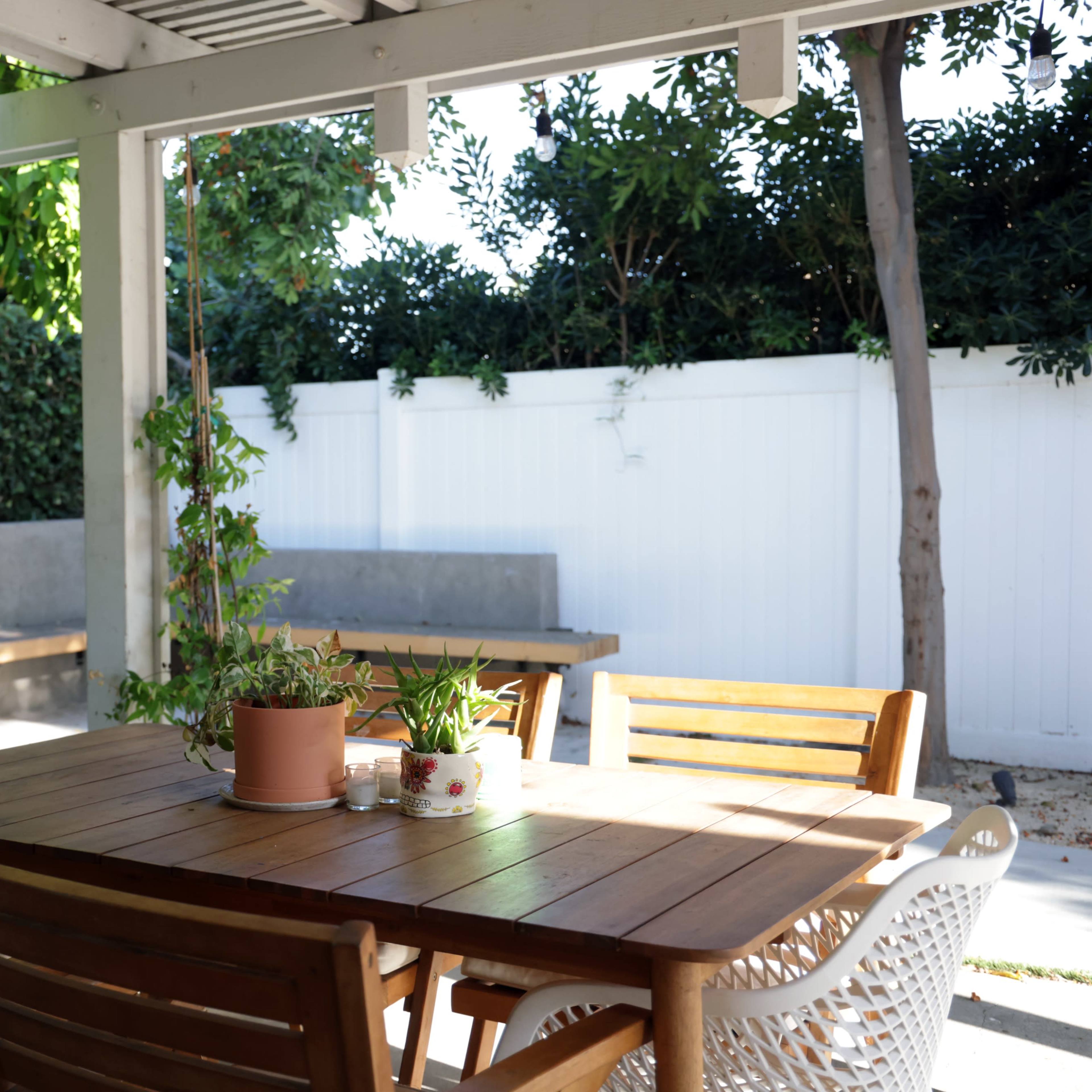 A wooden dining table with chairs is set under a covered patio, surrounded by greenery and a white fence.