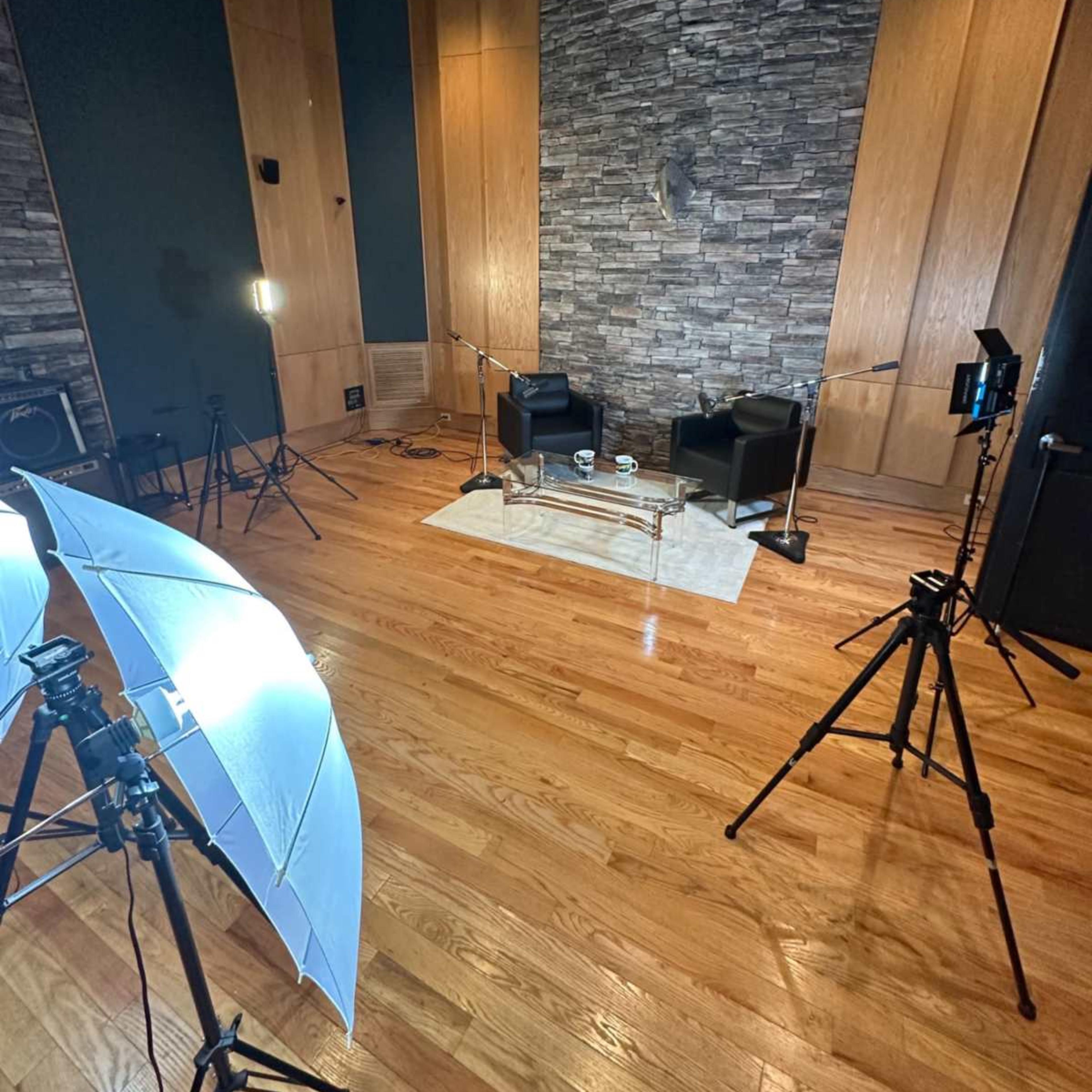A well-lit studio setting featuring two black chairs, a glass table, and multiple lighting equipment arranged around a wooden floor.