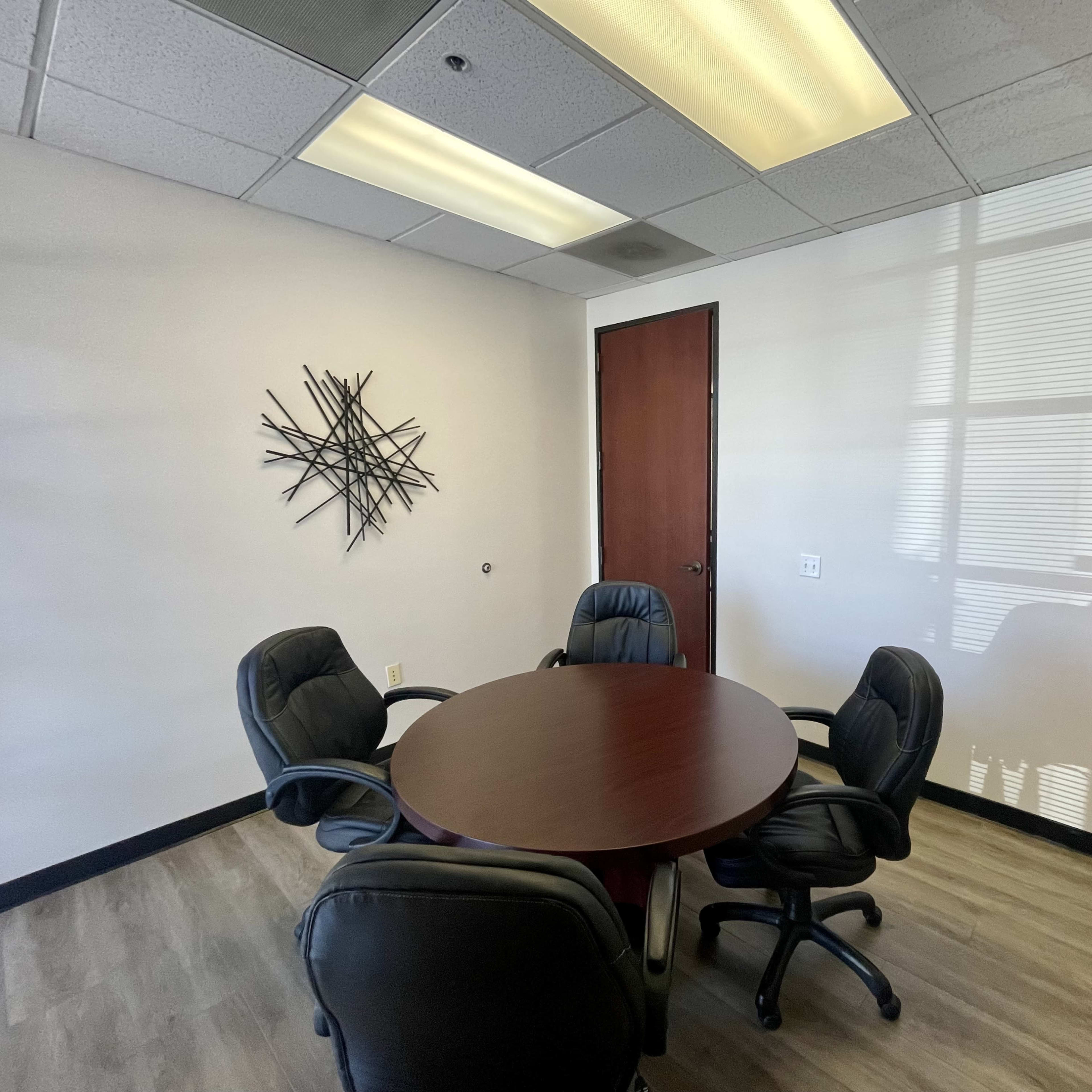 A small conference room features a round table surrounded by four black leather chairs, with a wall art piece and a wooden door.