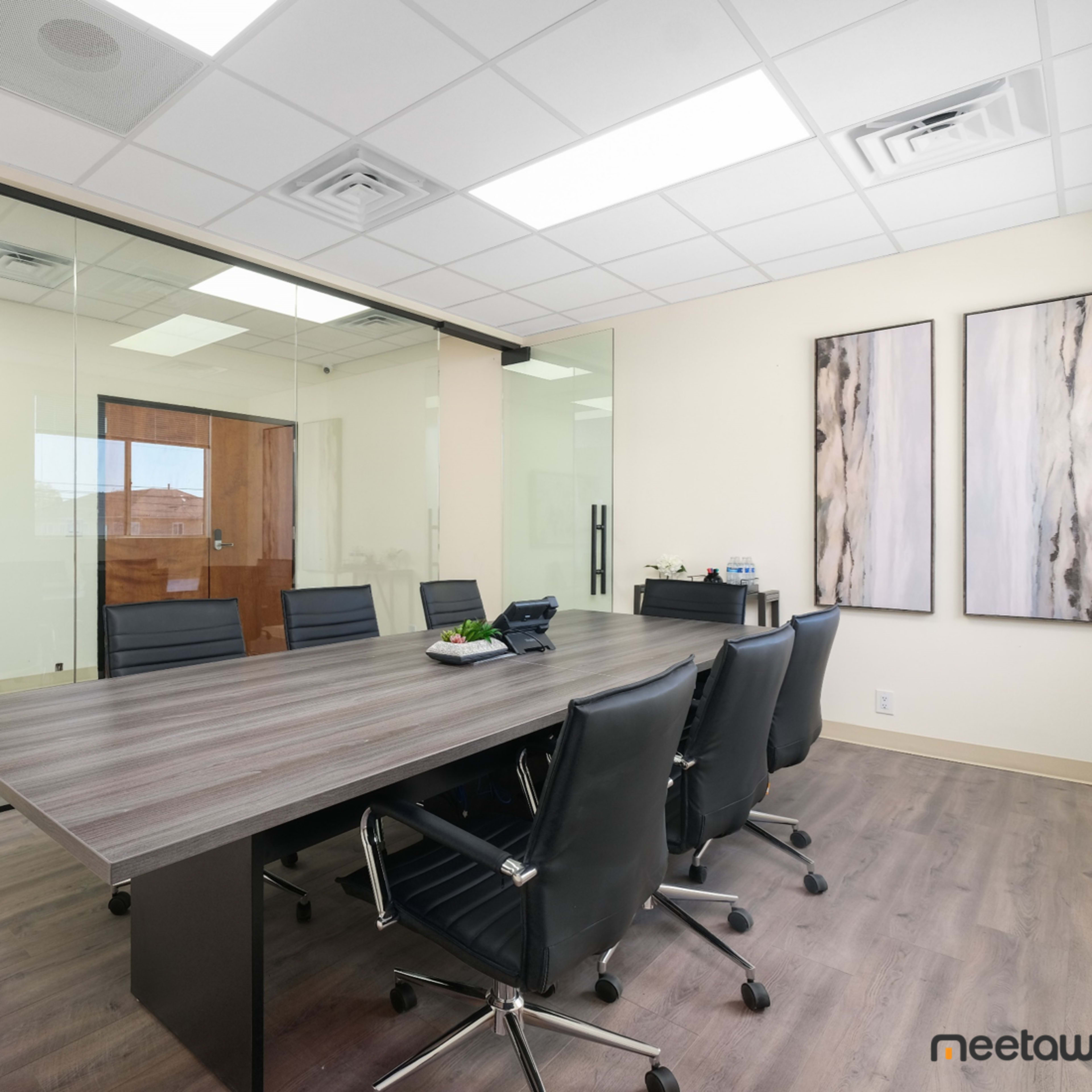 A modern conference room features a large wooden table surrounded by black chairs, with glass walls and artwork on the nearby walls.
