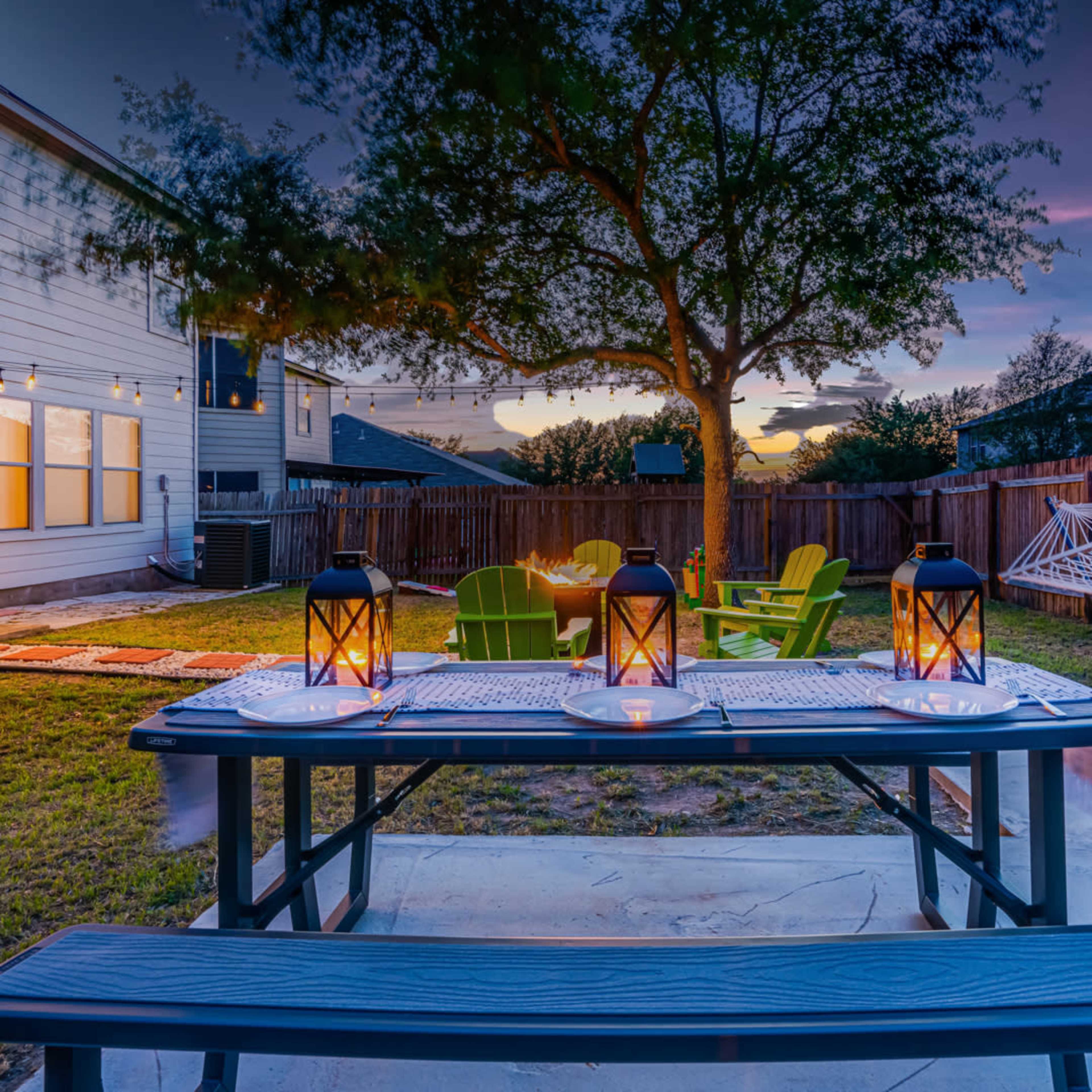 A backyard scene features a picnic table with lanterns, green chairs, a hammock, and a tree, set against a sunset sky.