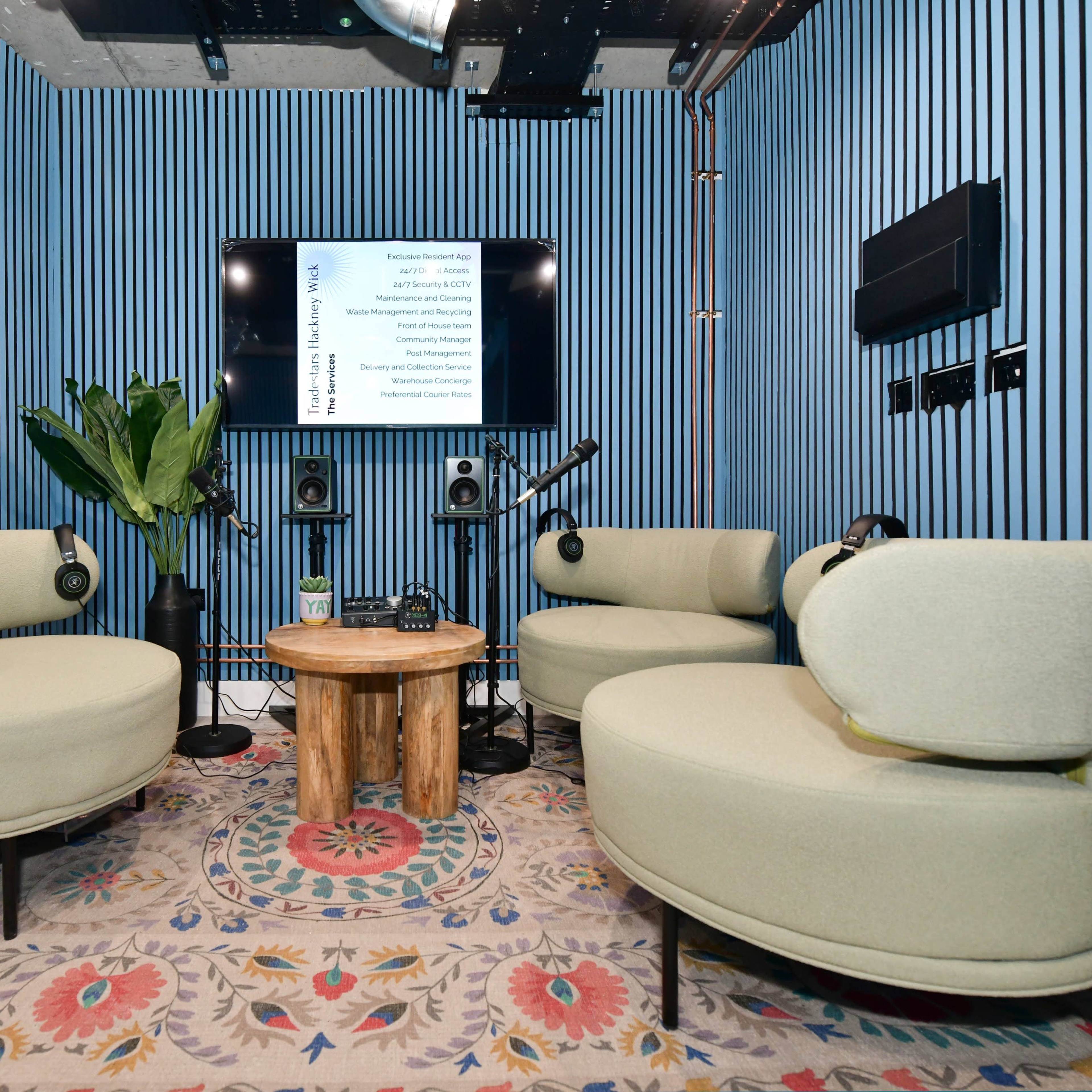 A small recording studio with two curved green couches, a wooden coffee table, and a screen displaying text, all set against blue striped walls.