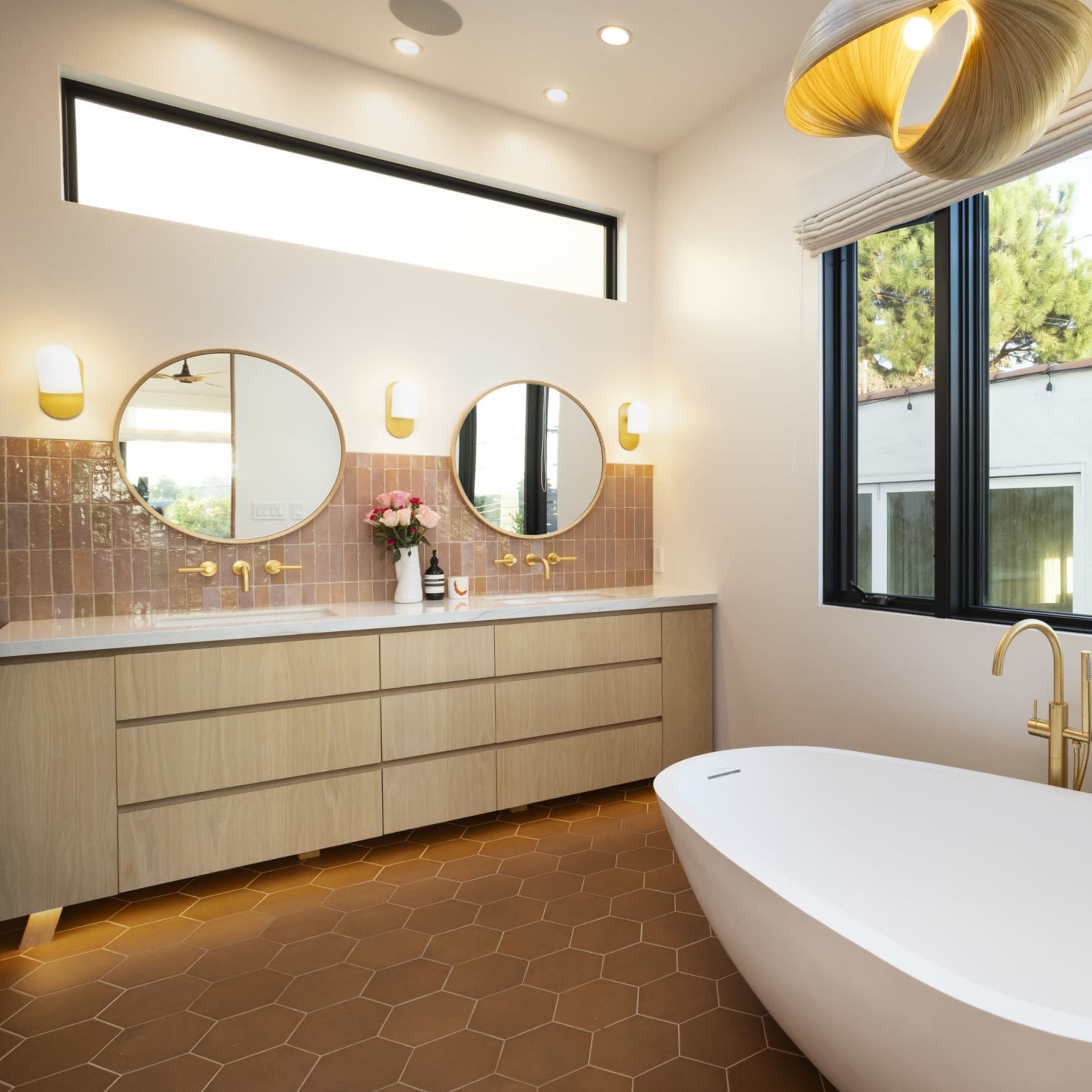 A modern bathroom features a freestanding bathtub, dual round mirrors, and a wooden vanity with gold fixtures, illuminated by natural light from large windows.