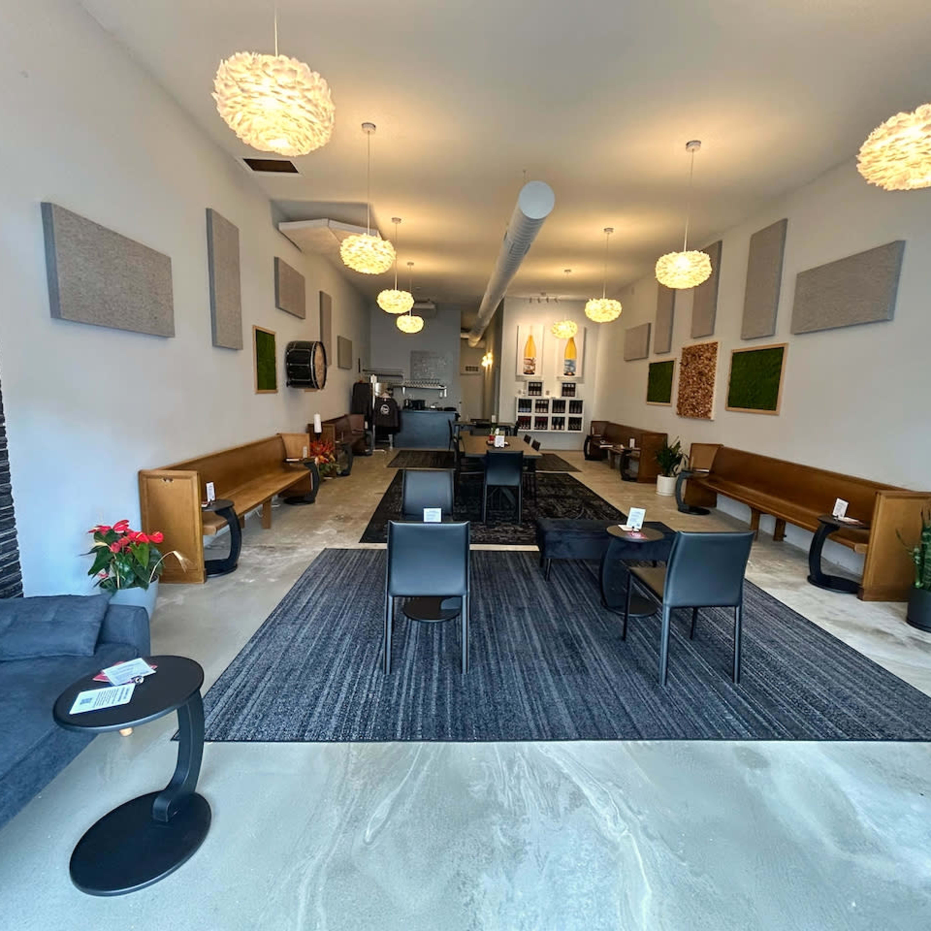 A spacious interior of a minimally decorated lounge with wooden benches, black chairs, and plants, along with pendant lighting and sound-absorbing panels on the walls.