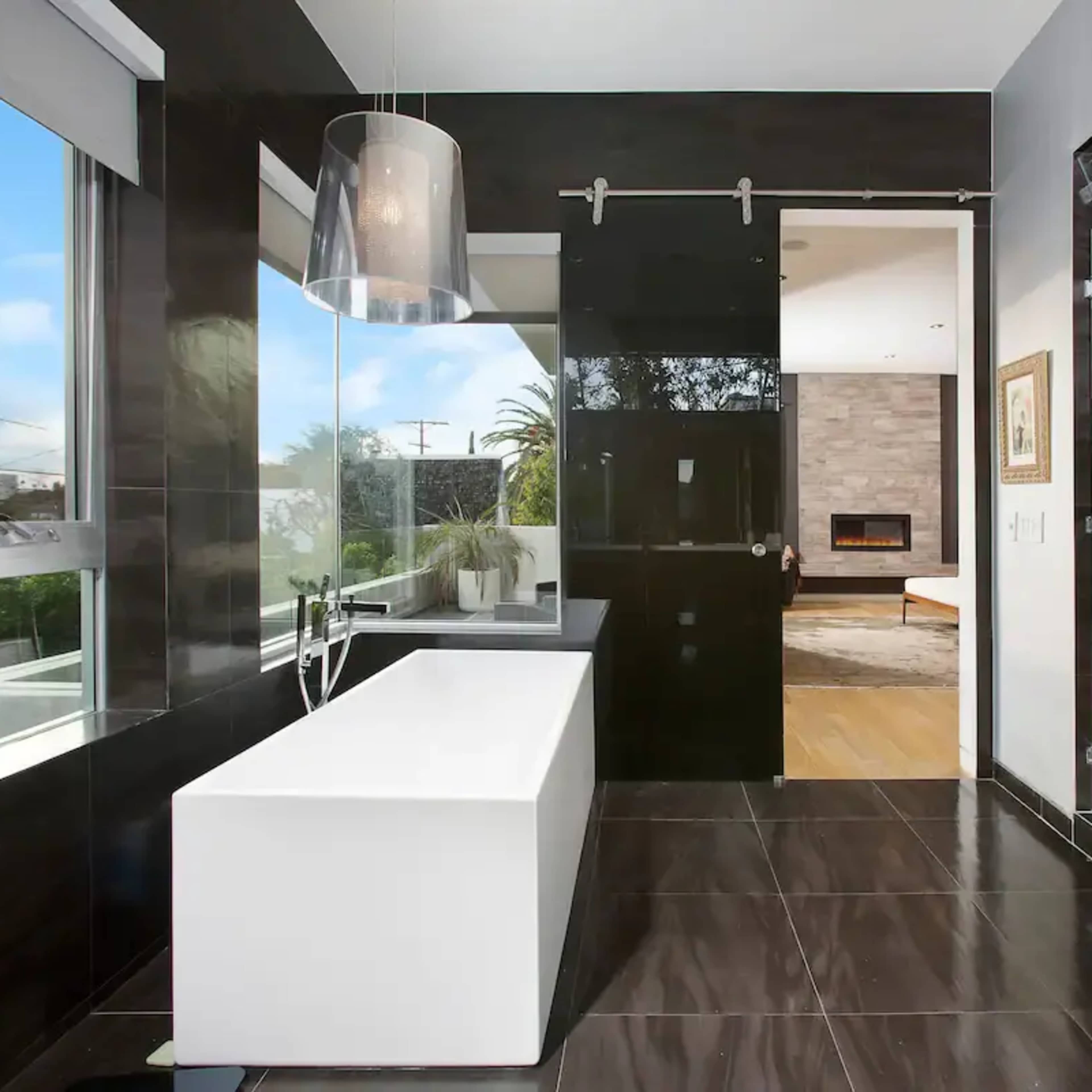 A modern bathroom with a freestanding white bathtub, a glass shower enclosure, and dark tiled walls and flooring.