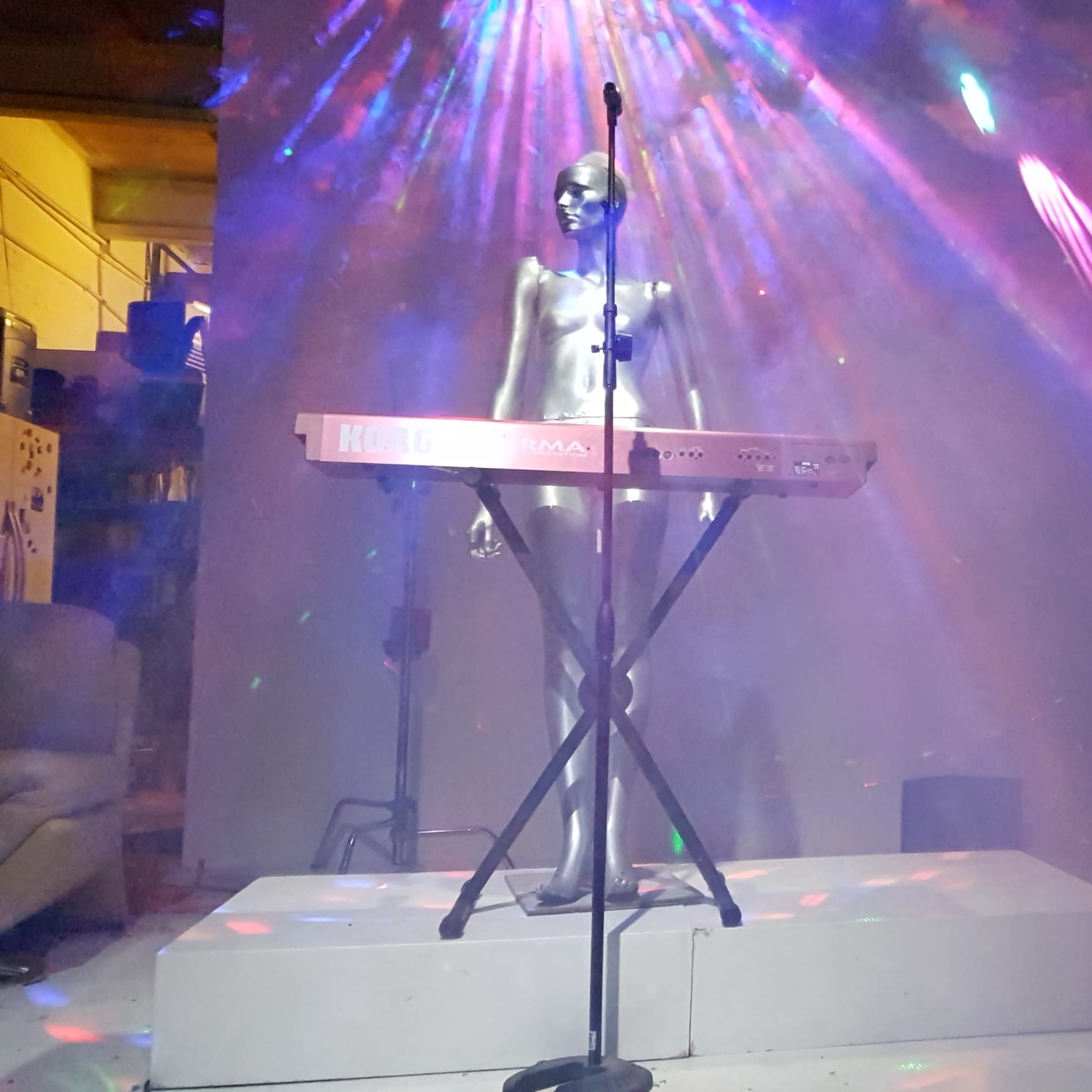 A silver mannequin stands behind a keyboard on a stage illuminated by colorful laser lights and smoke.