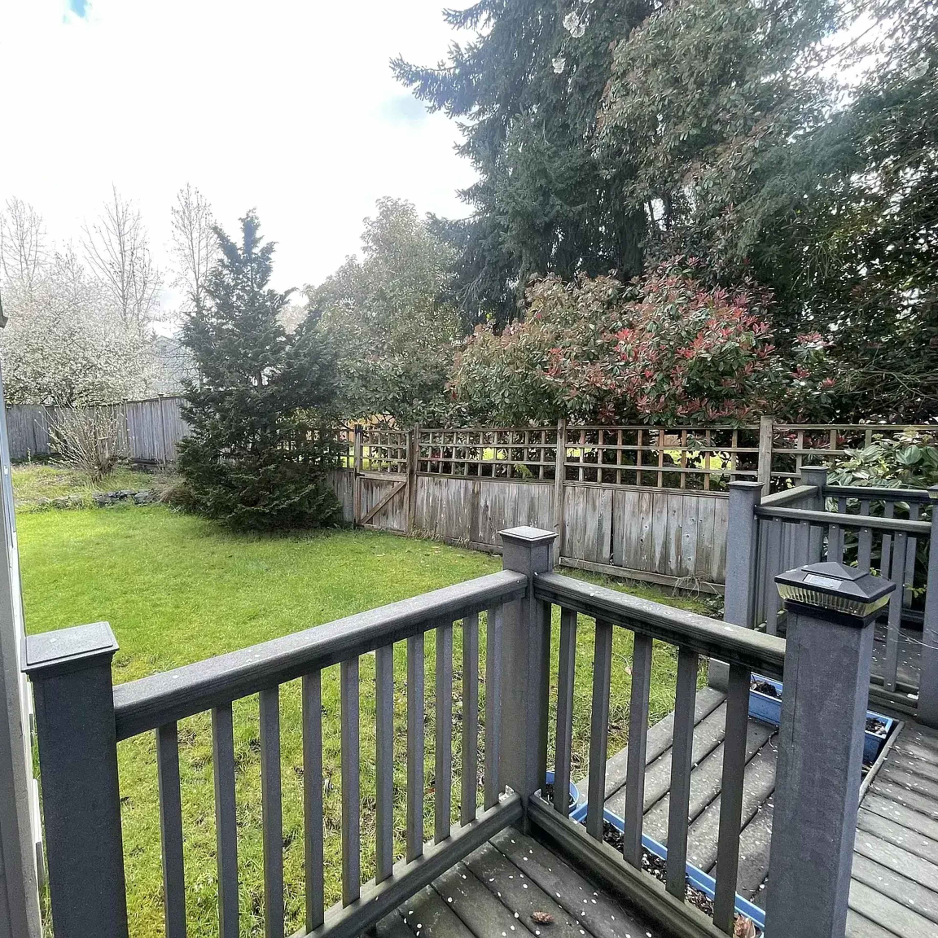A wooden deck overlooks a grassy backyard surrounded by trees and a wooden fence.