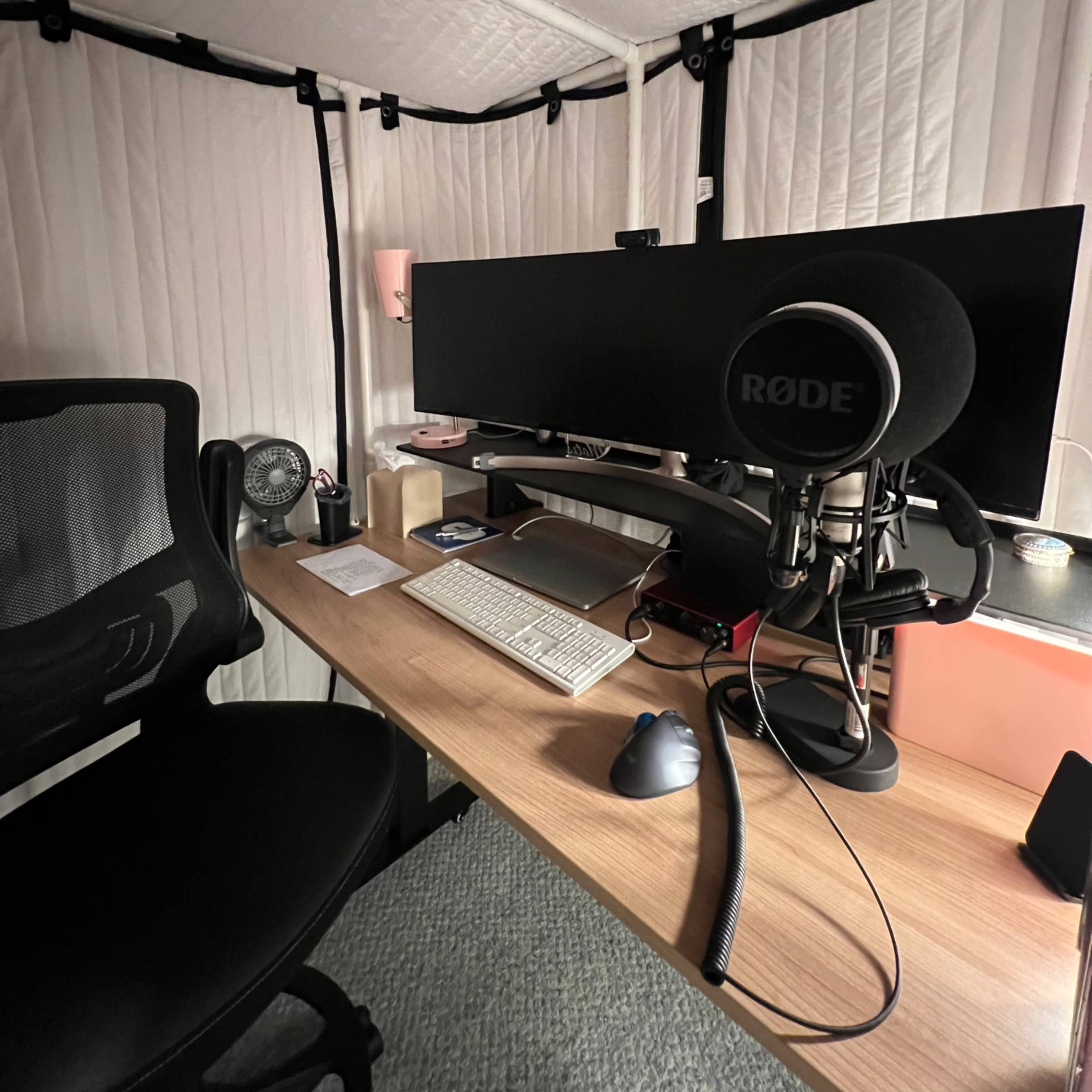 A desk with a computer monitor, microphone, keyboard, and mouse inside a soundproofed booth.