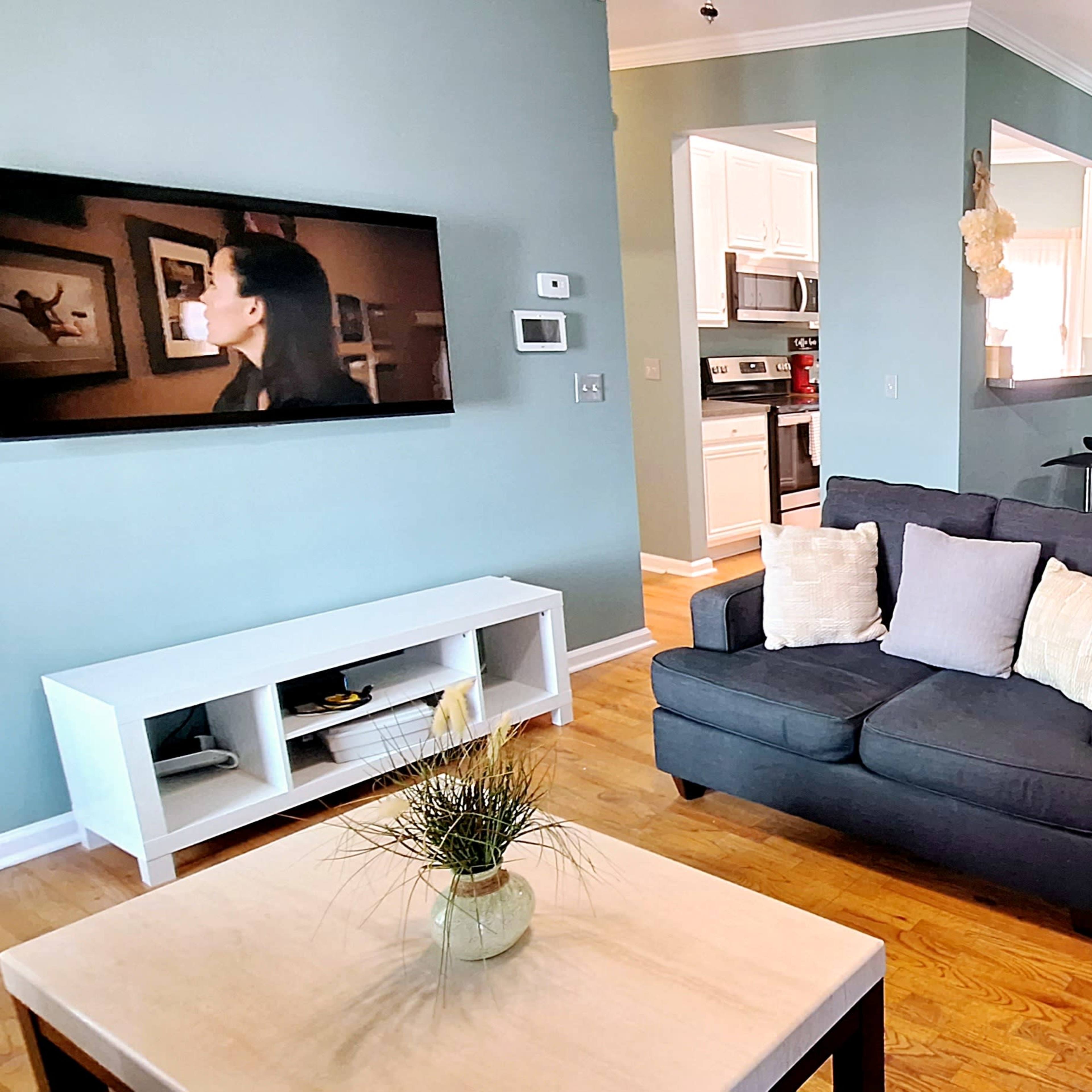 A living room with a dark sofa facing a television, adjacent to a dining area and a kitchen visible in the background.