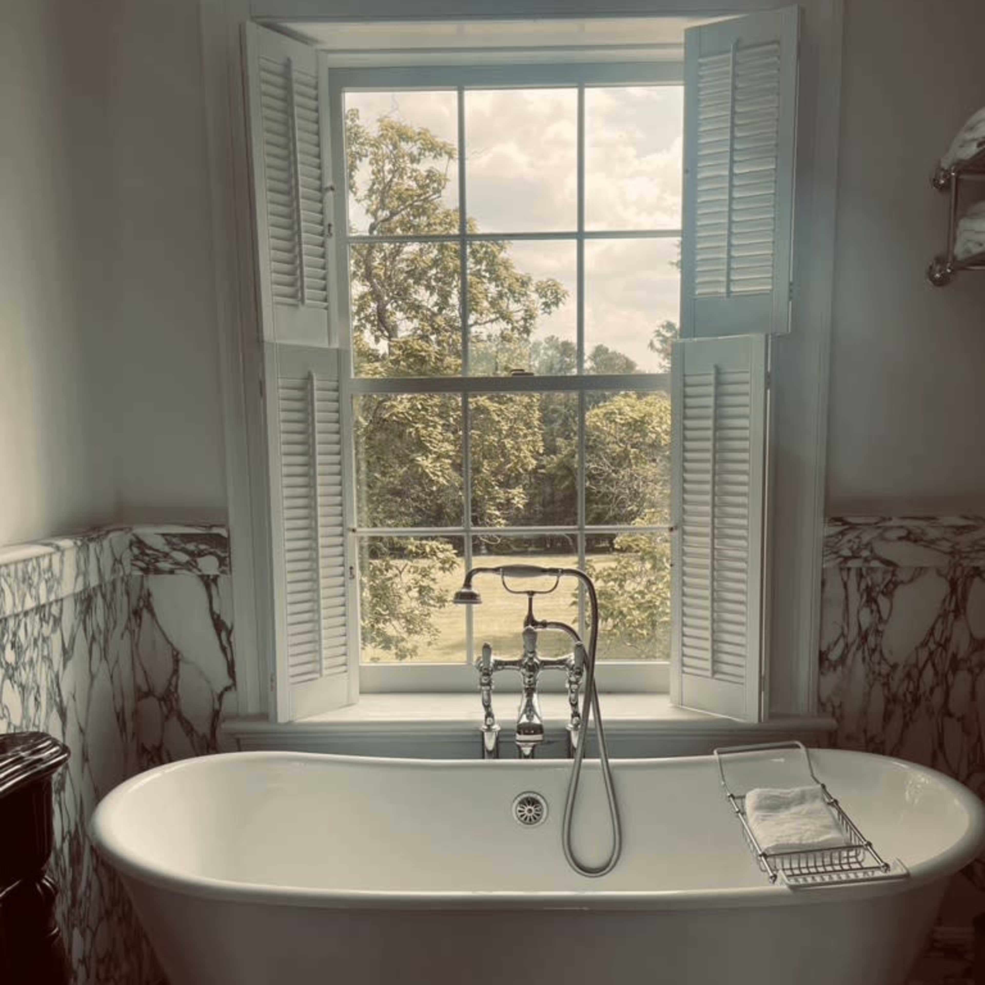A freestanding bathtub is positioned in front of a large window with shutters, offering a view of trees outside.