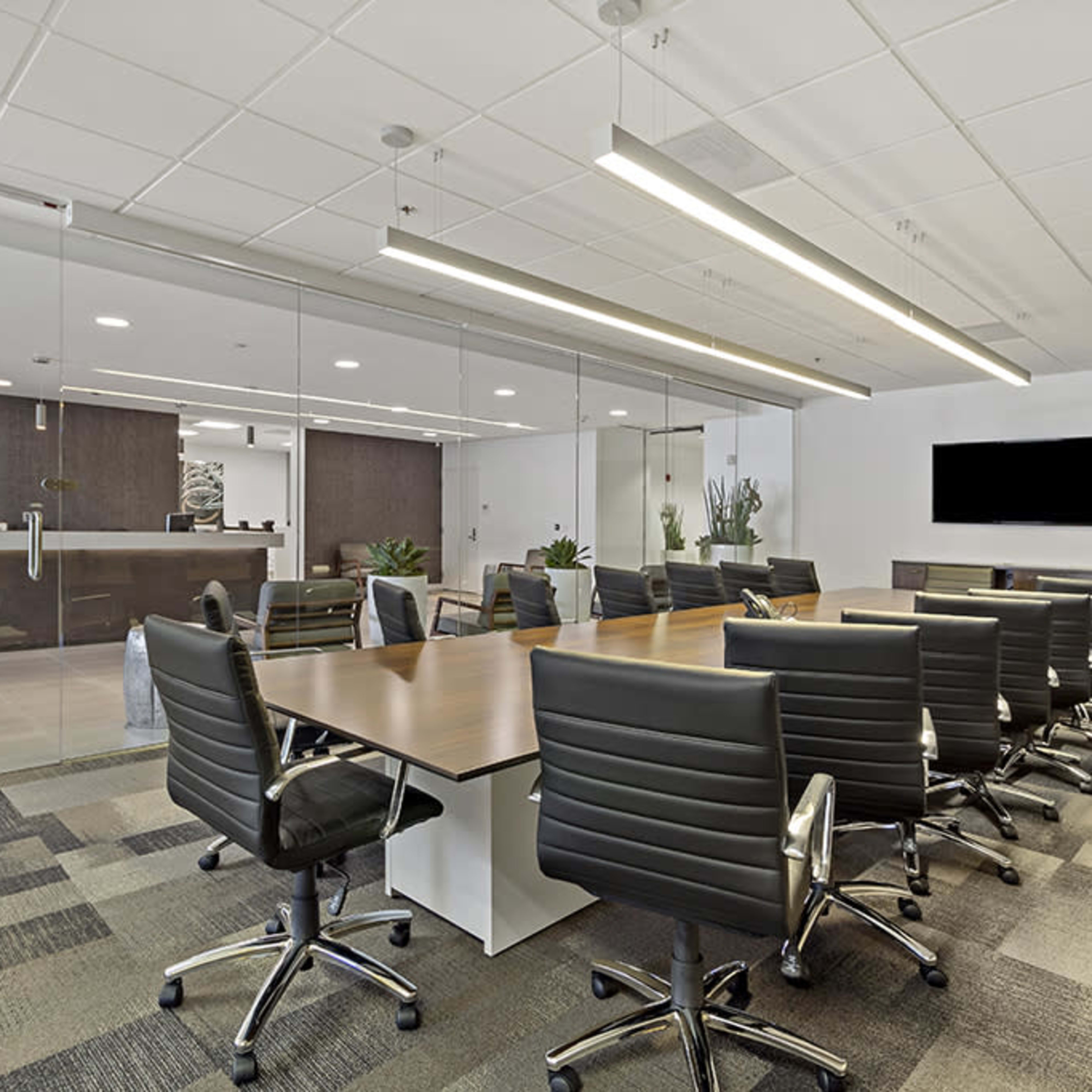 A modern conference room features a long wooden table surrounded by black rolling chairs, with a glass wall overlooking a reception area.
