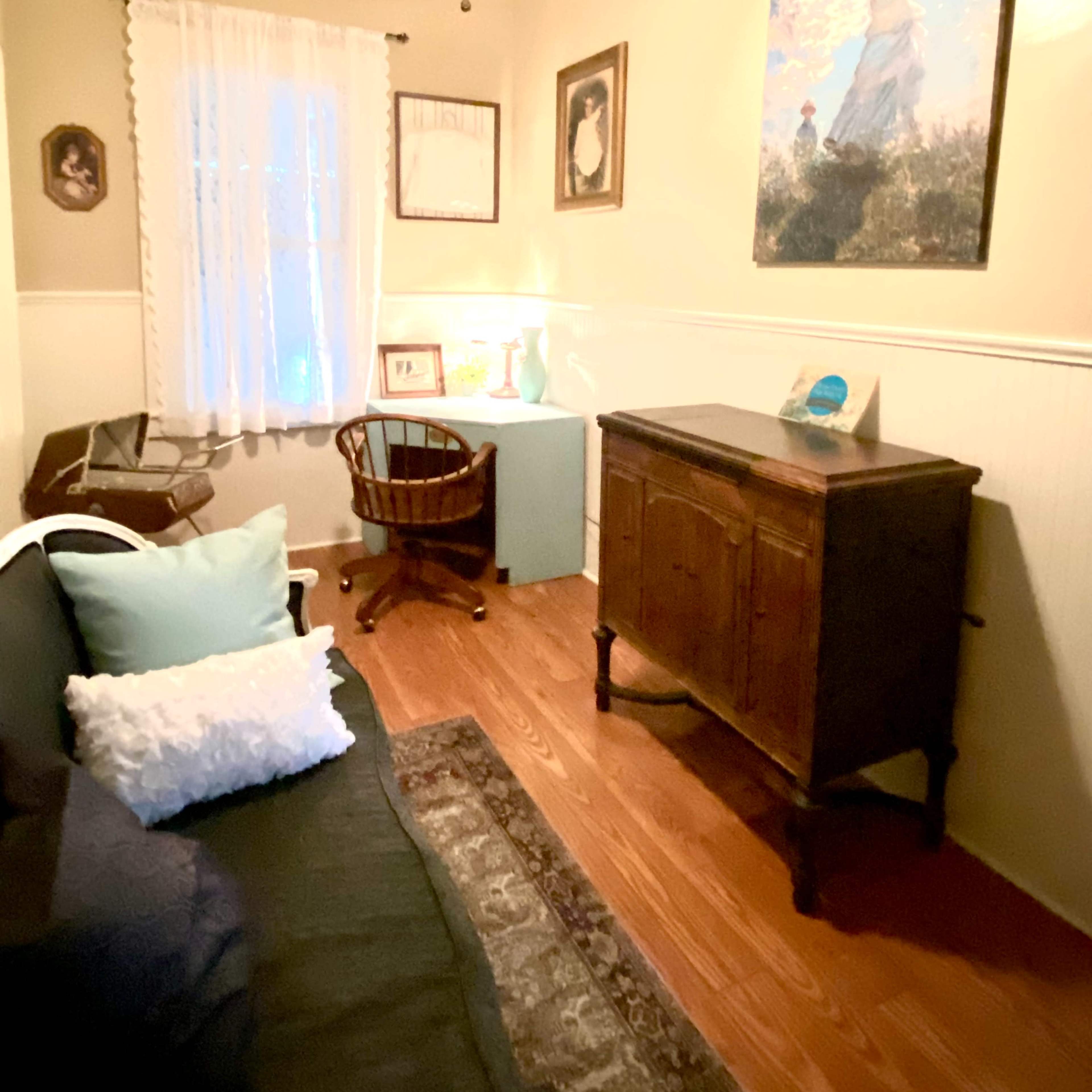 A small room with a wooden floor, a ceiling fan, a wall-mounted painting, and a cozy seating area including a black couch and a wooden chair.