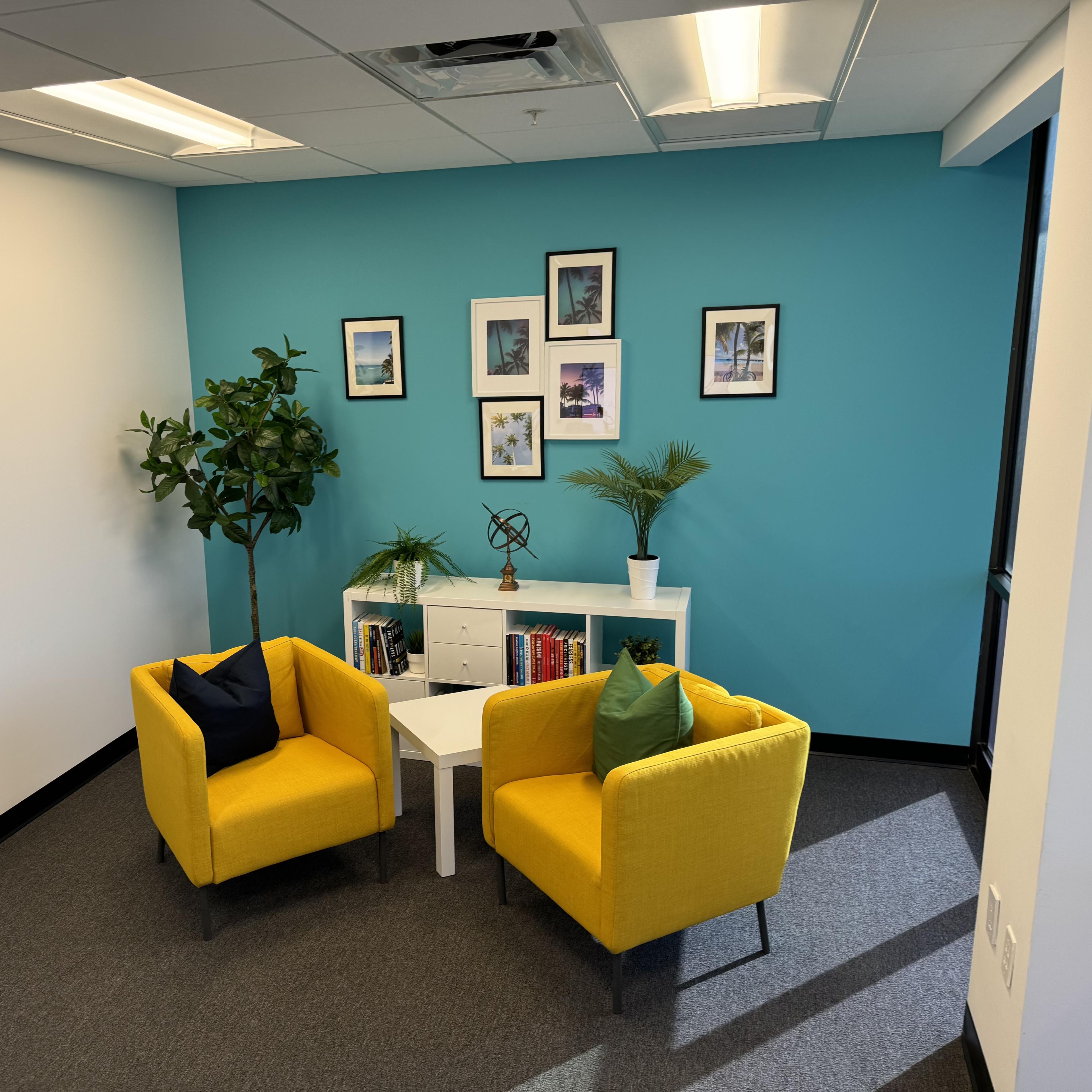 A small, modern seating area with two yellow chairs, a white coffee table, and decorative plants against a blue wall adorned with framed pictures.