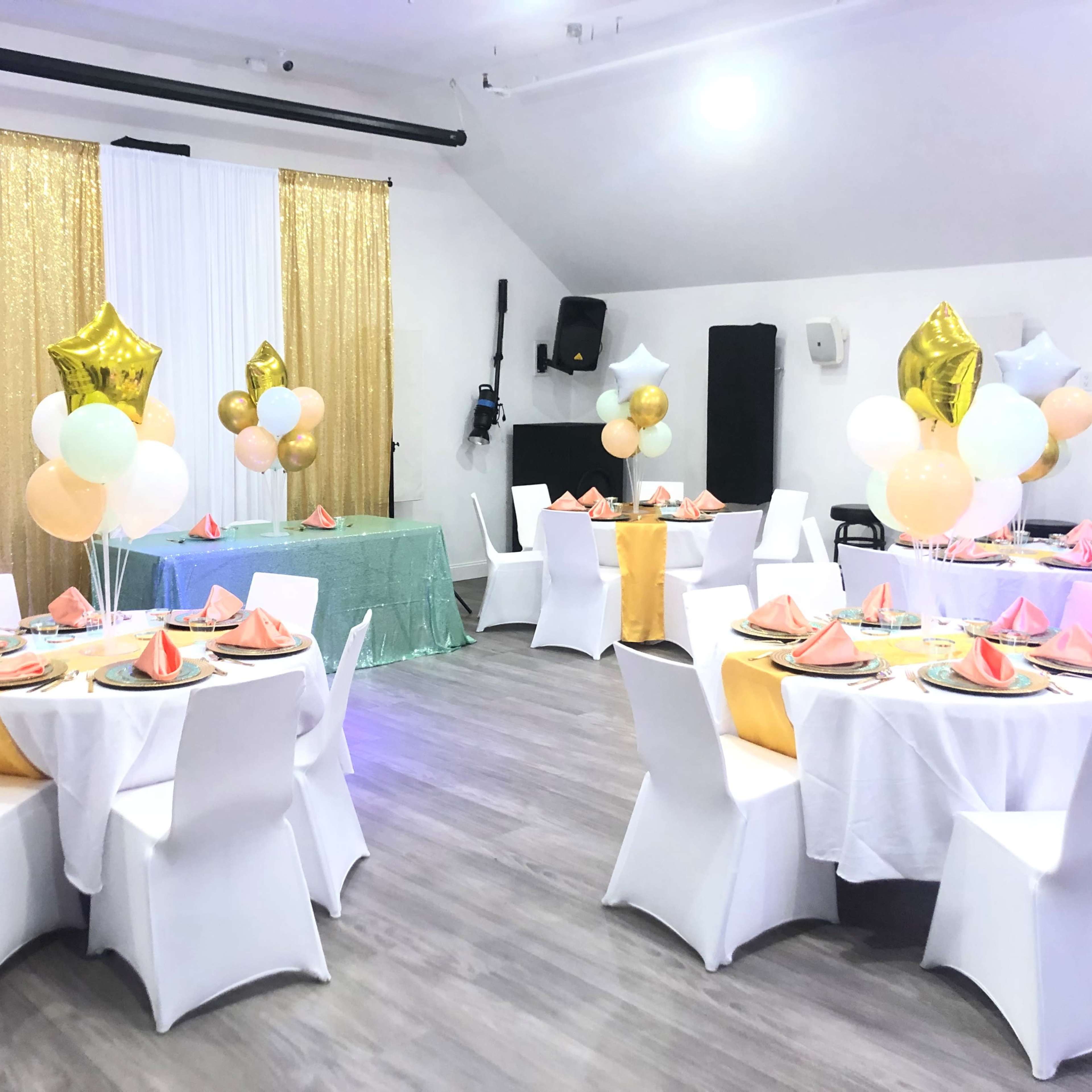 A decorated event space featuring white tables with colorful table settings, gold and green accents, and balloon arrangements.
