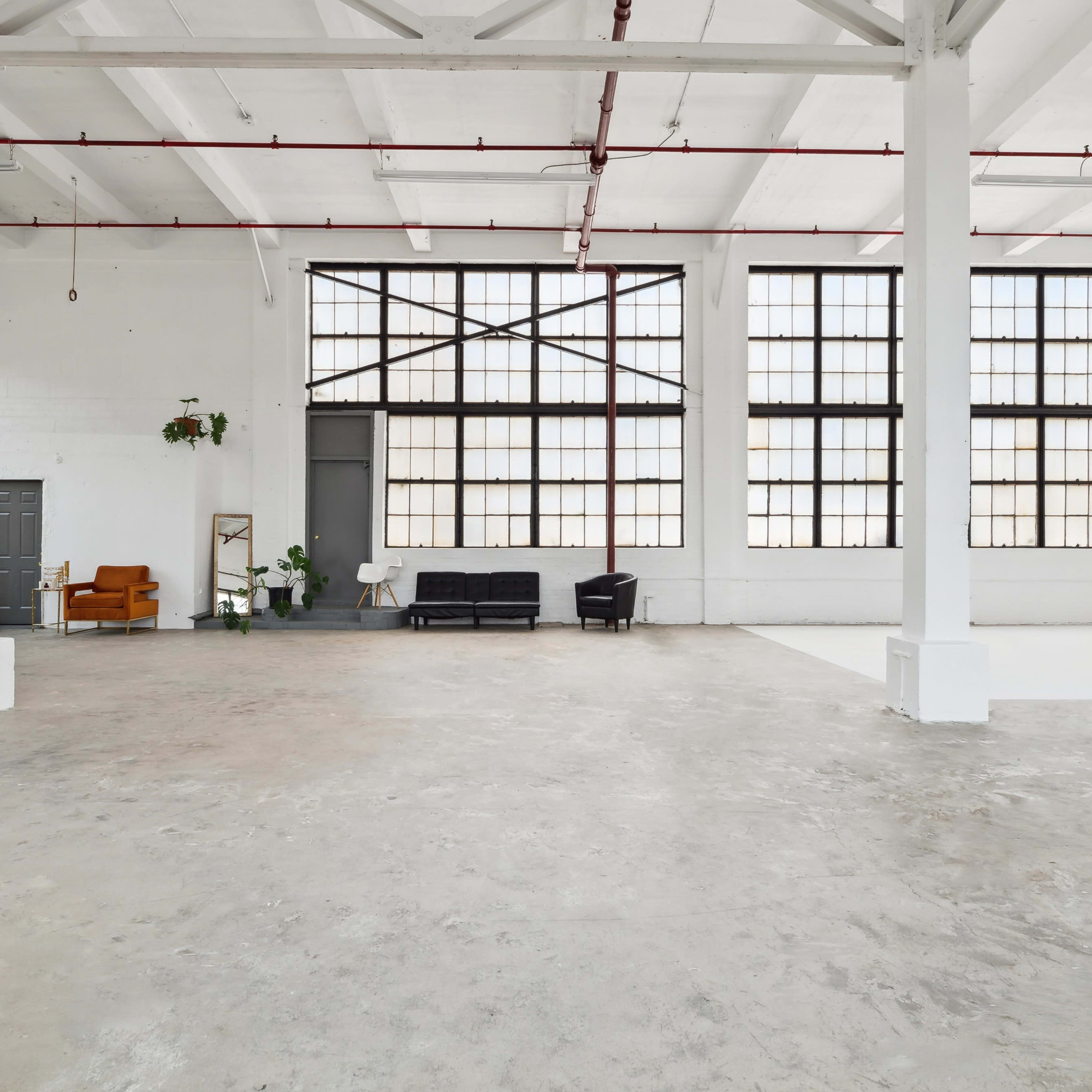 A spacious, industrial-style interior with large windows, bare concrete floors, and minimalist furniture arranged in a well-lit environment.