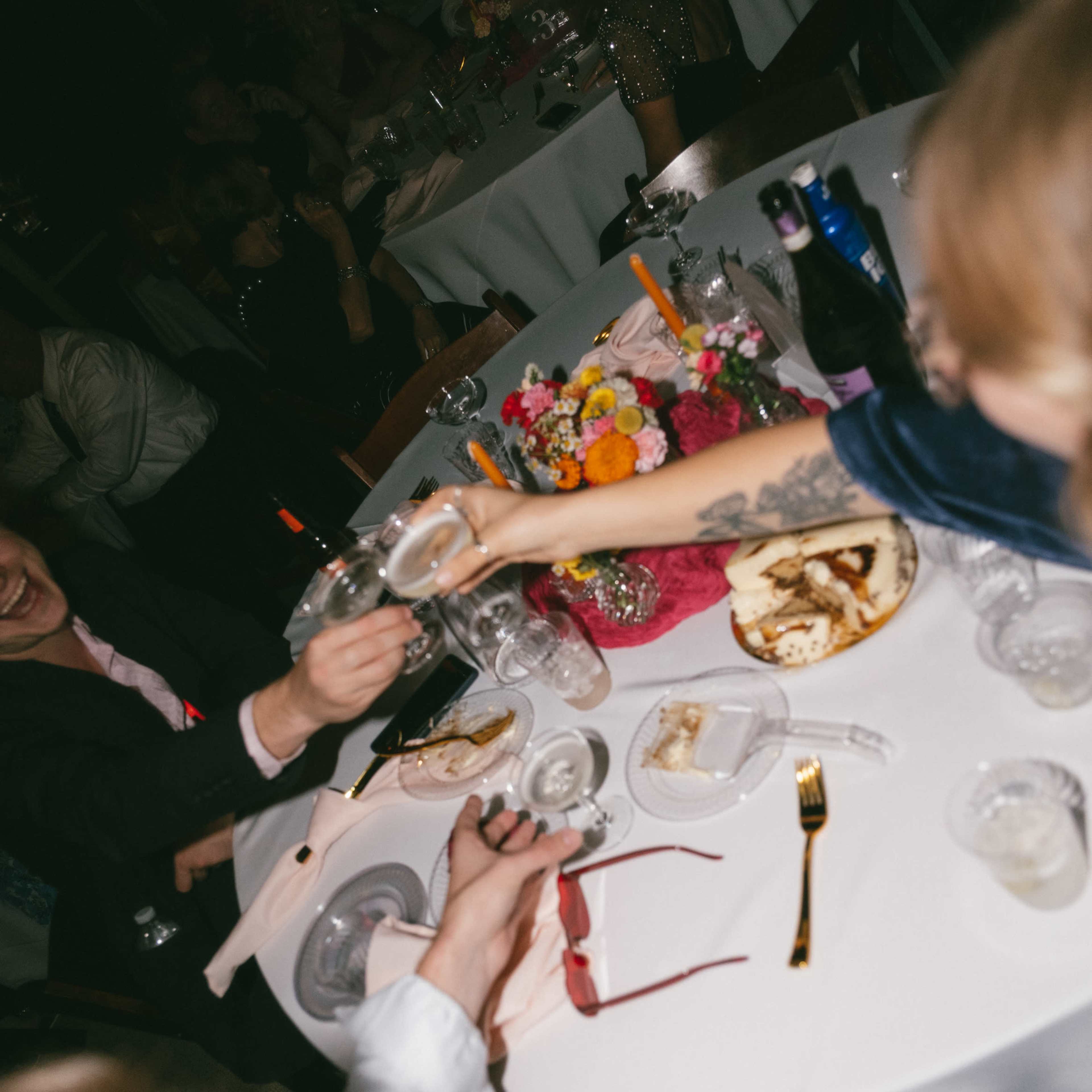 People at a dinner table are toasting with glasses amid plates of food and floral arrangements.