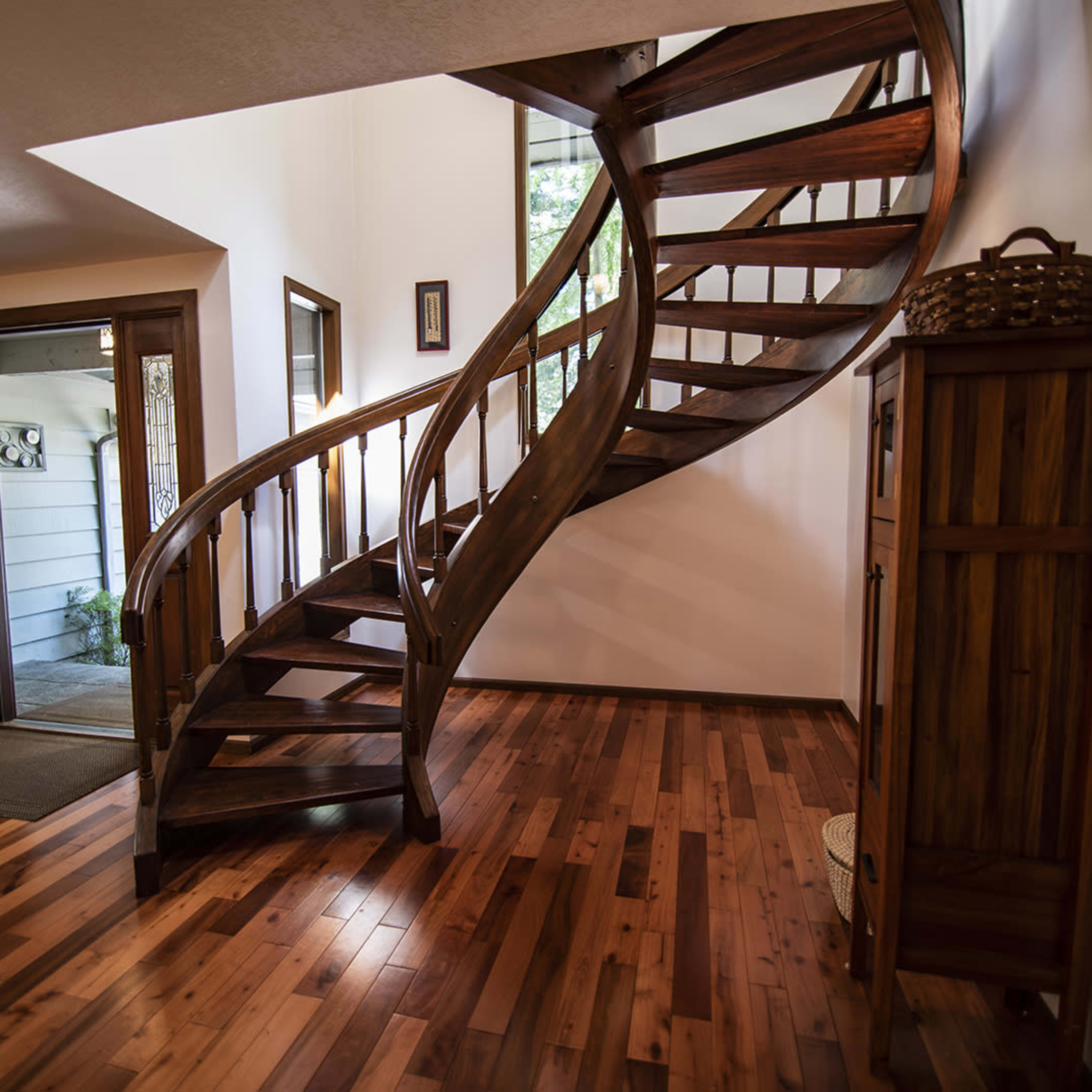 A wooden spiral staircase leading to an upper level, set within a warmly lit entryway that includes a front door and a wooden cabinet.