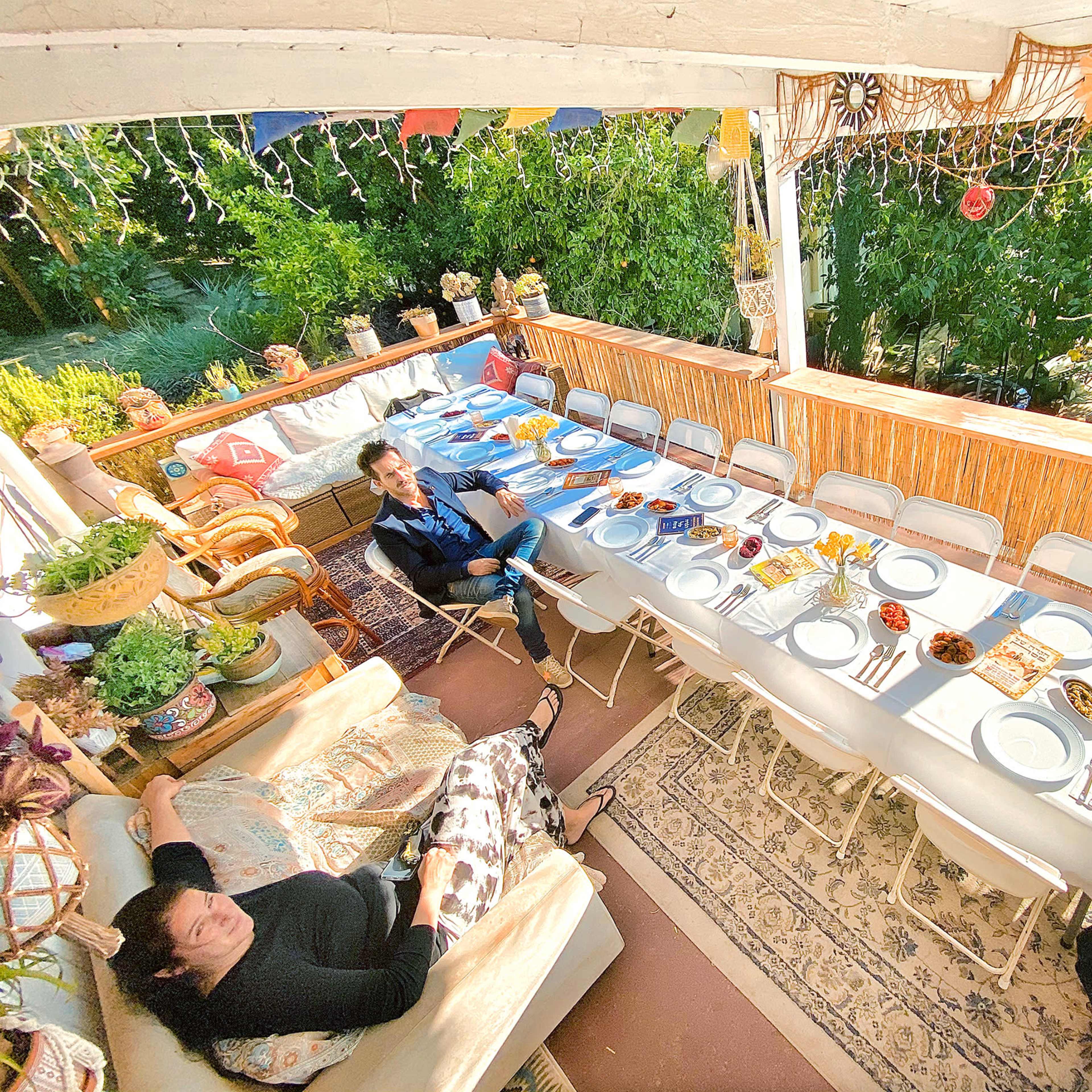 A long table is set for a meal on a patio, with chairs arranged around it and two people seated nearby, surrounded by greenery and decorative items.