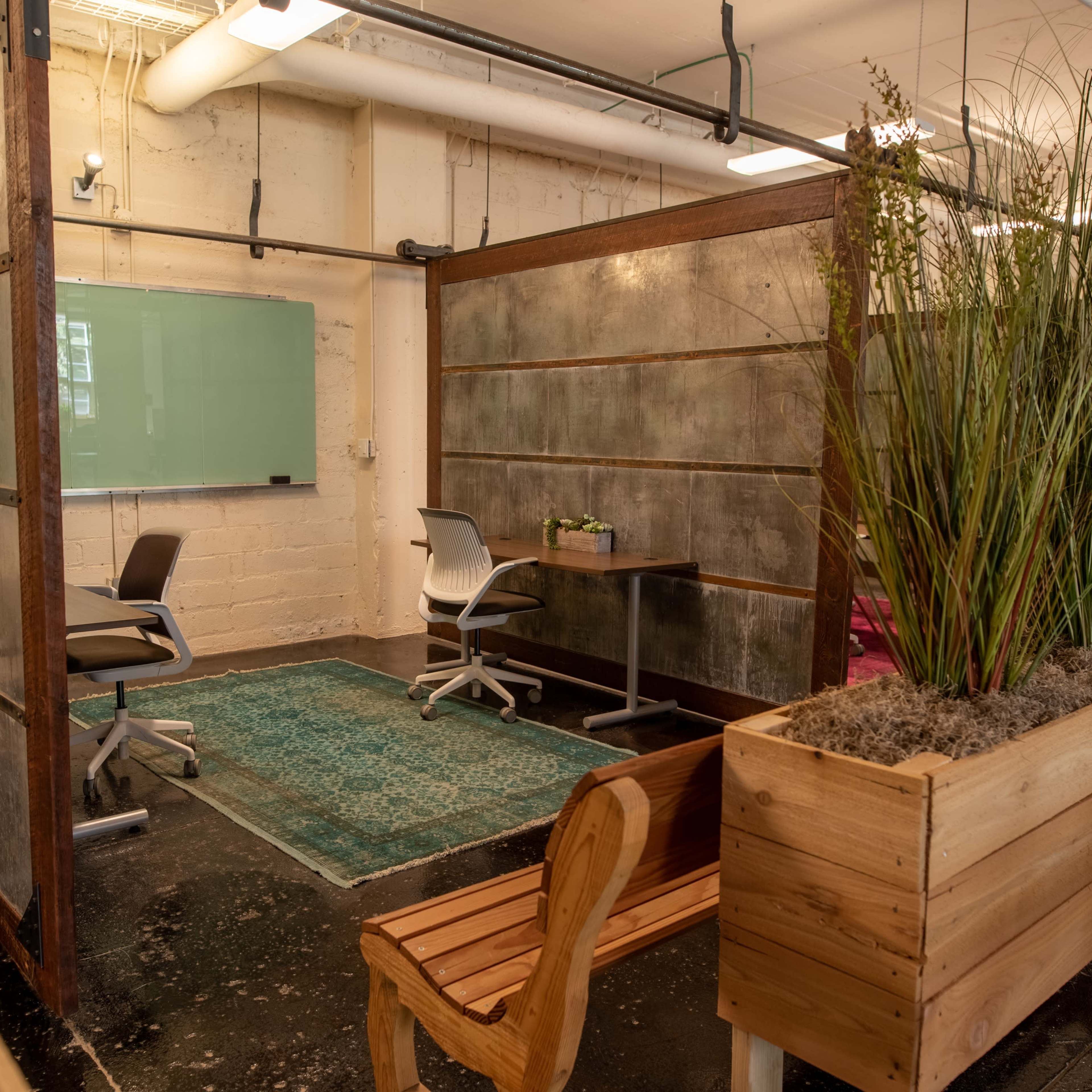 A modern workspace with partitioned areas featuring a rug, chairs, a table, and planter boxes with greenery.