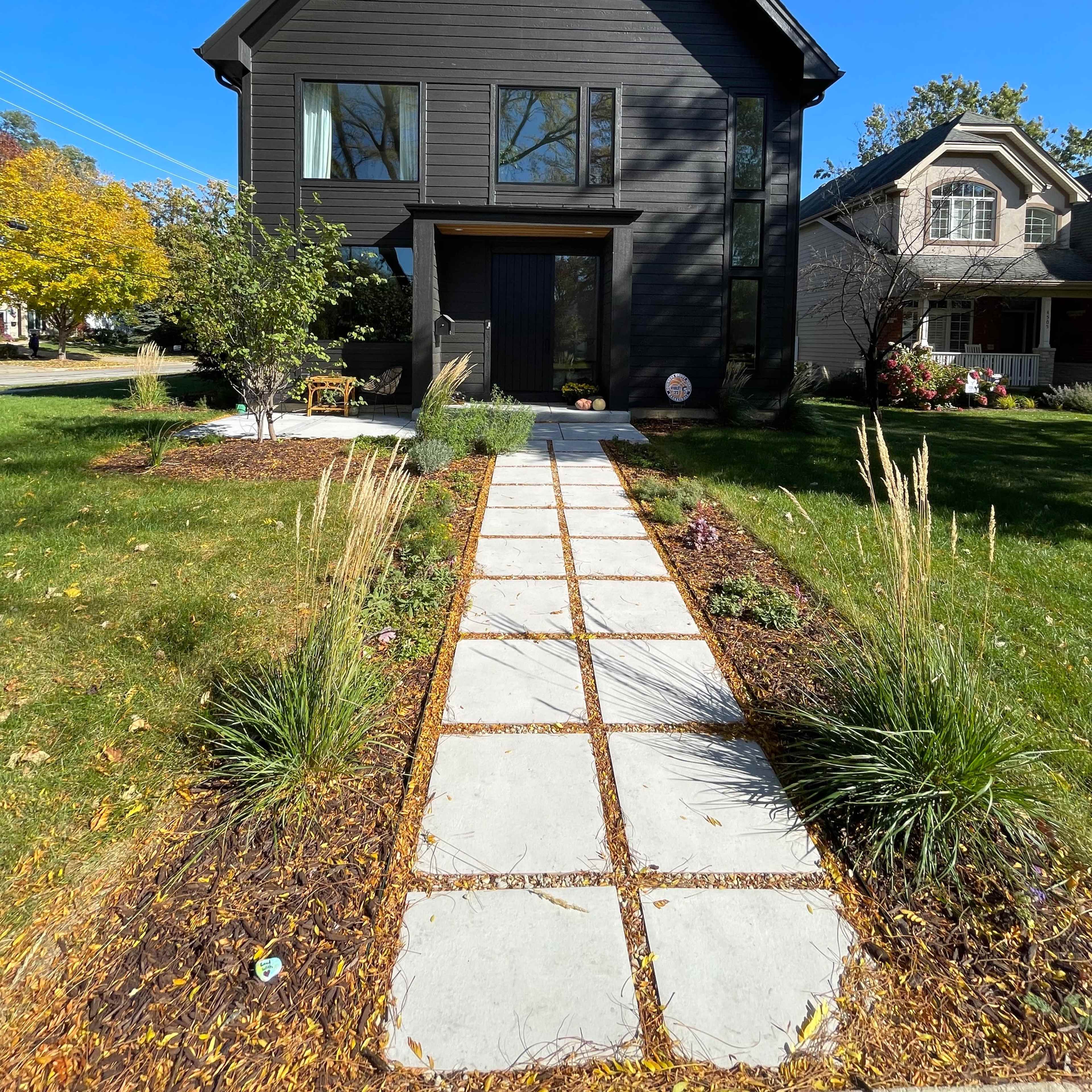 A modern black house stands at the end of a stone pathway lined with grass and shrubs.