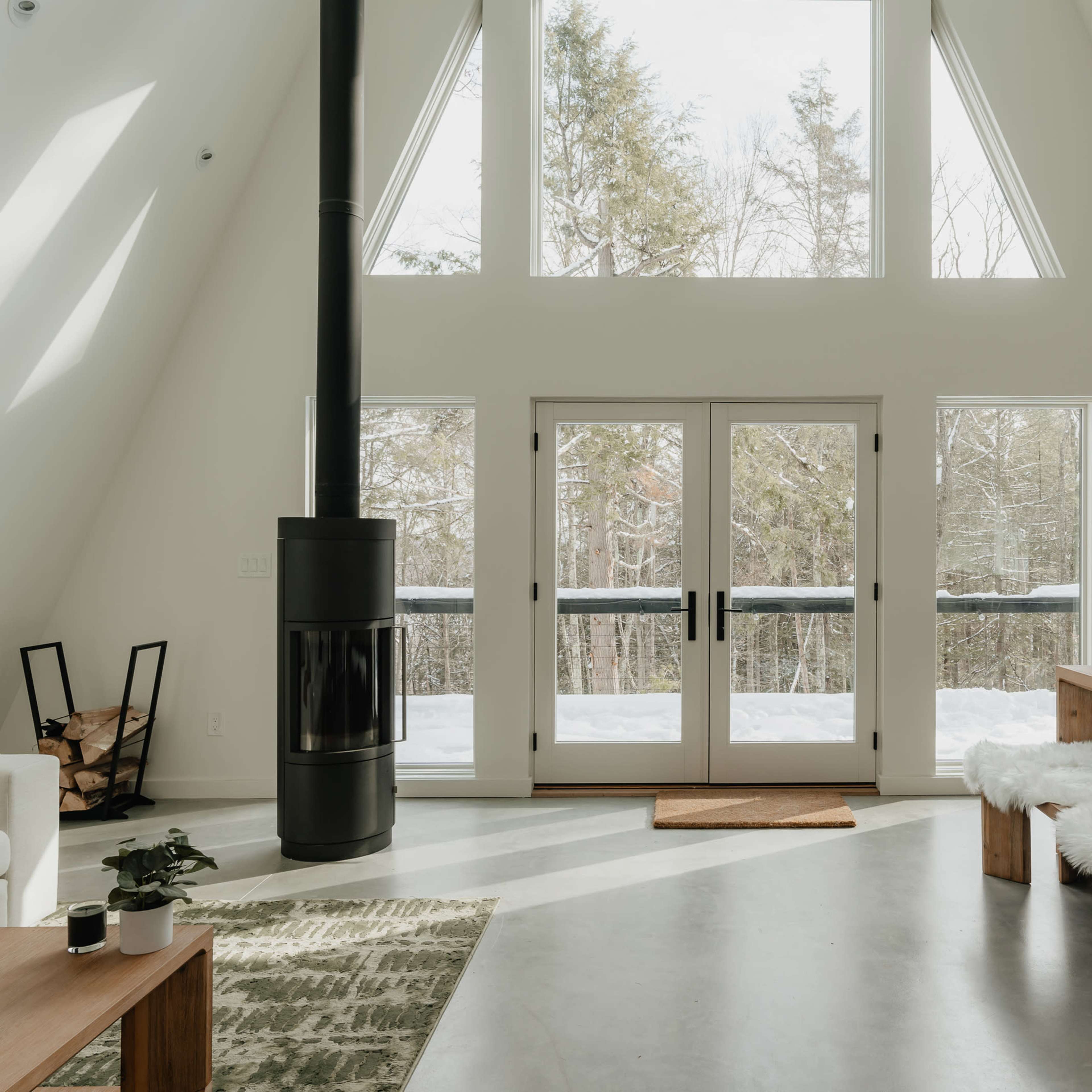 A bright, modern room features large windows, a wooden dining table, and a central wood stove, with snowy trees visible outside.