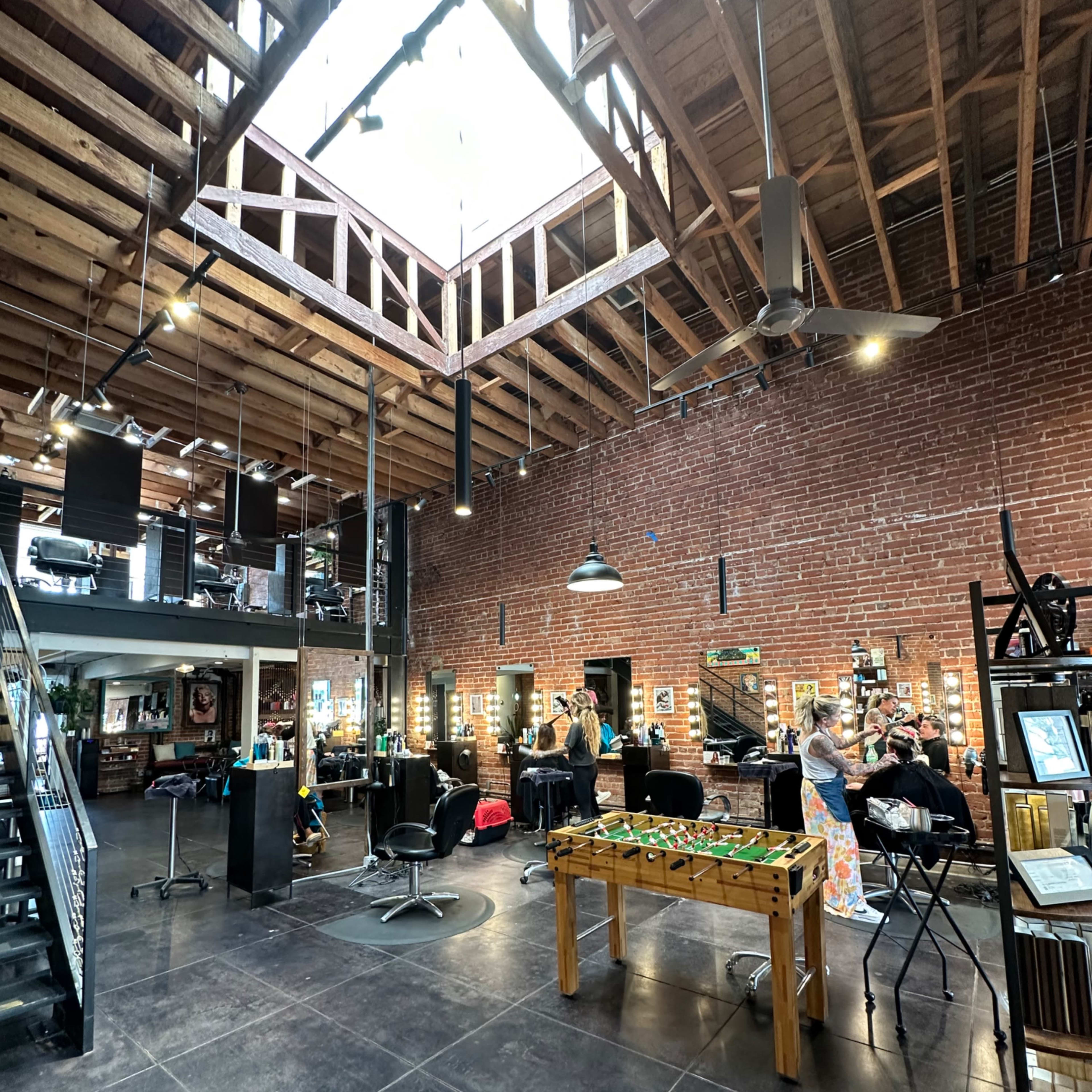A spacious barbershop with a high wooden ceiling, exposed brick walls, and several styling stations, including a foosball table in the center.