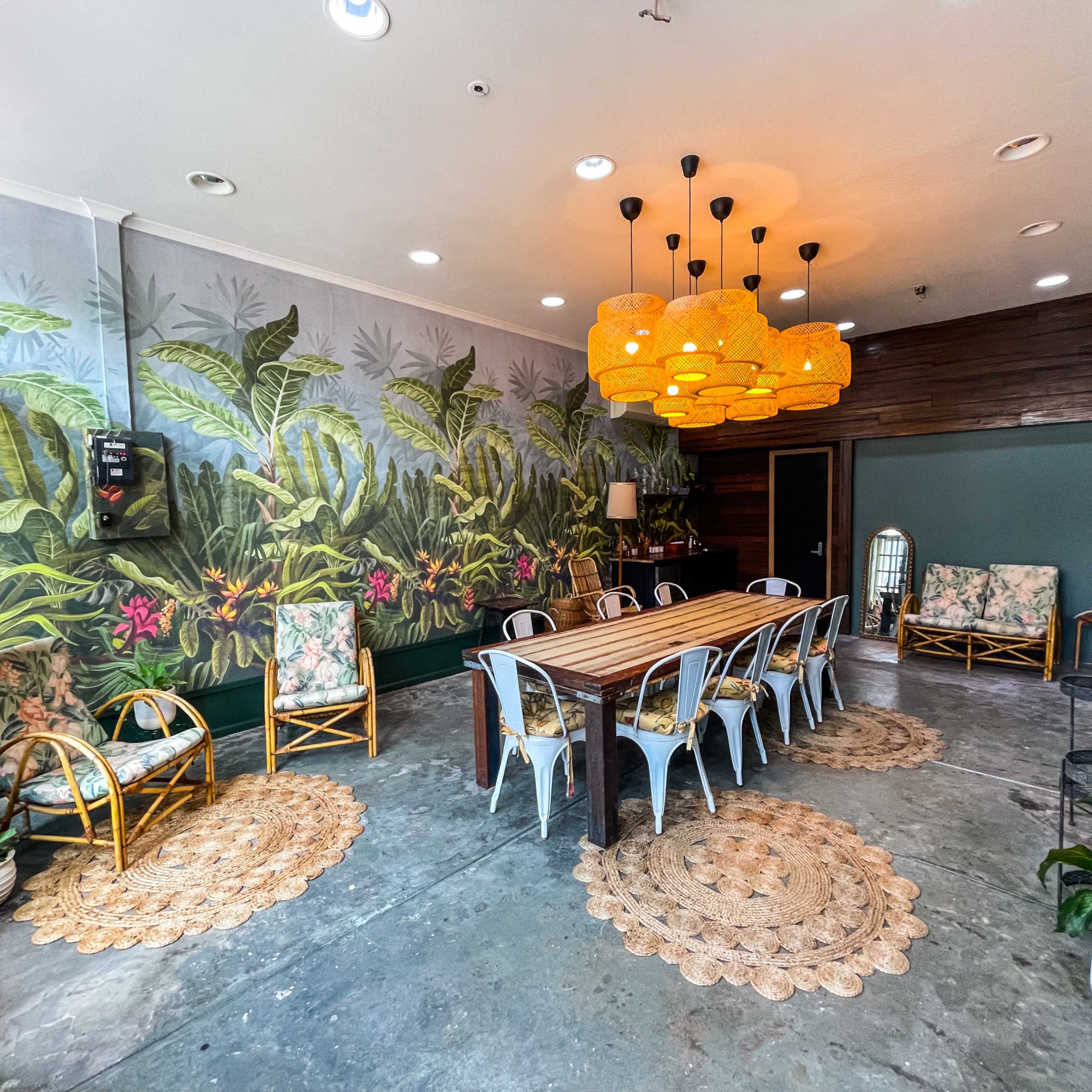 A dining area featuring a large wooden table surrounded by chairs and complemented by tropical-themed wallpaper, decorative lighting, and plants.
