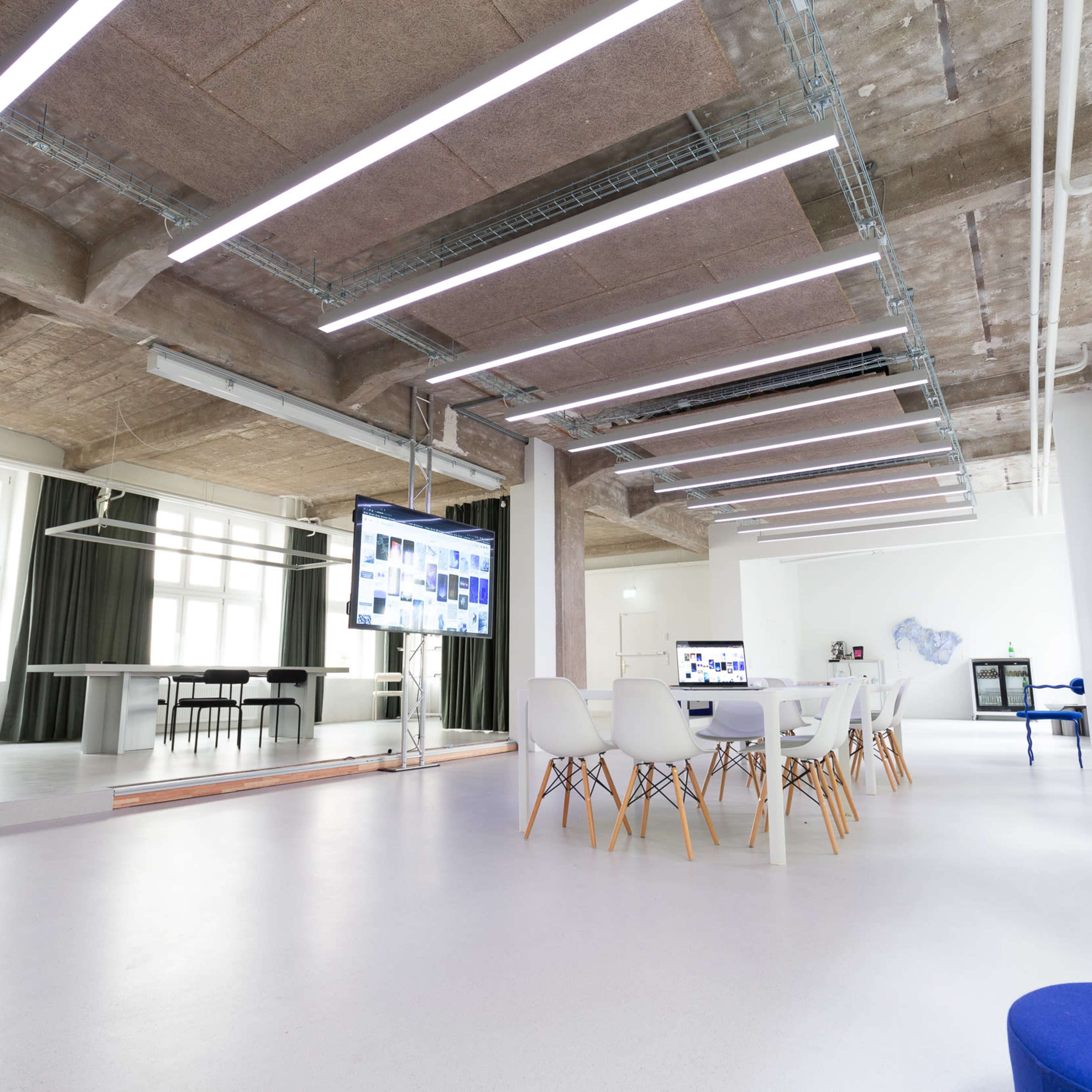 A modern office space with white walls, a large table surrounded by chairs, and a screen displaying digital content.