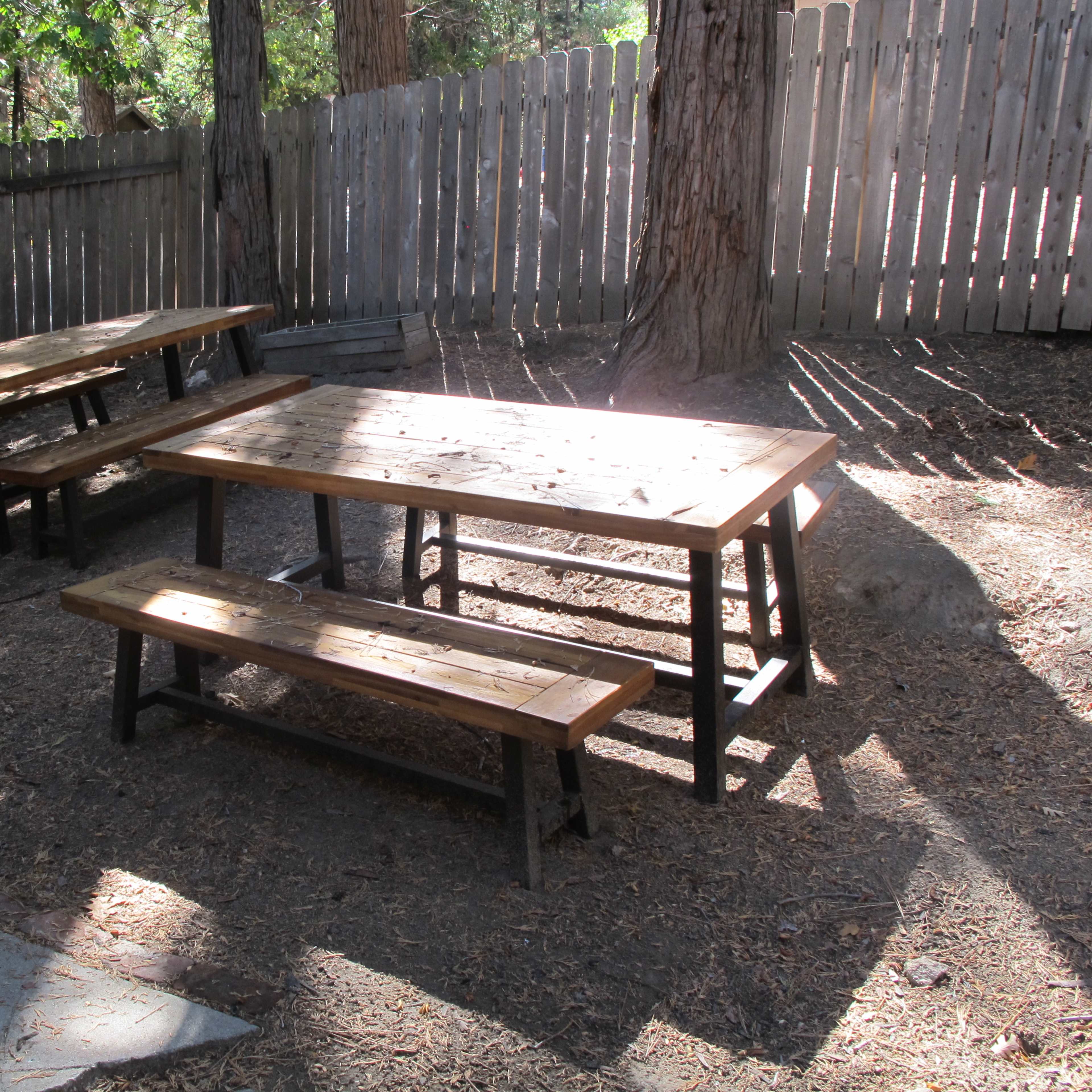 A wooden picnic table and benches in a shaded outdoor area surrounded by trees and a wooden fence.