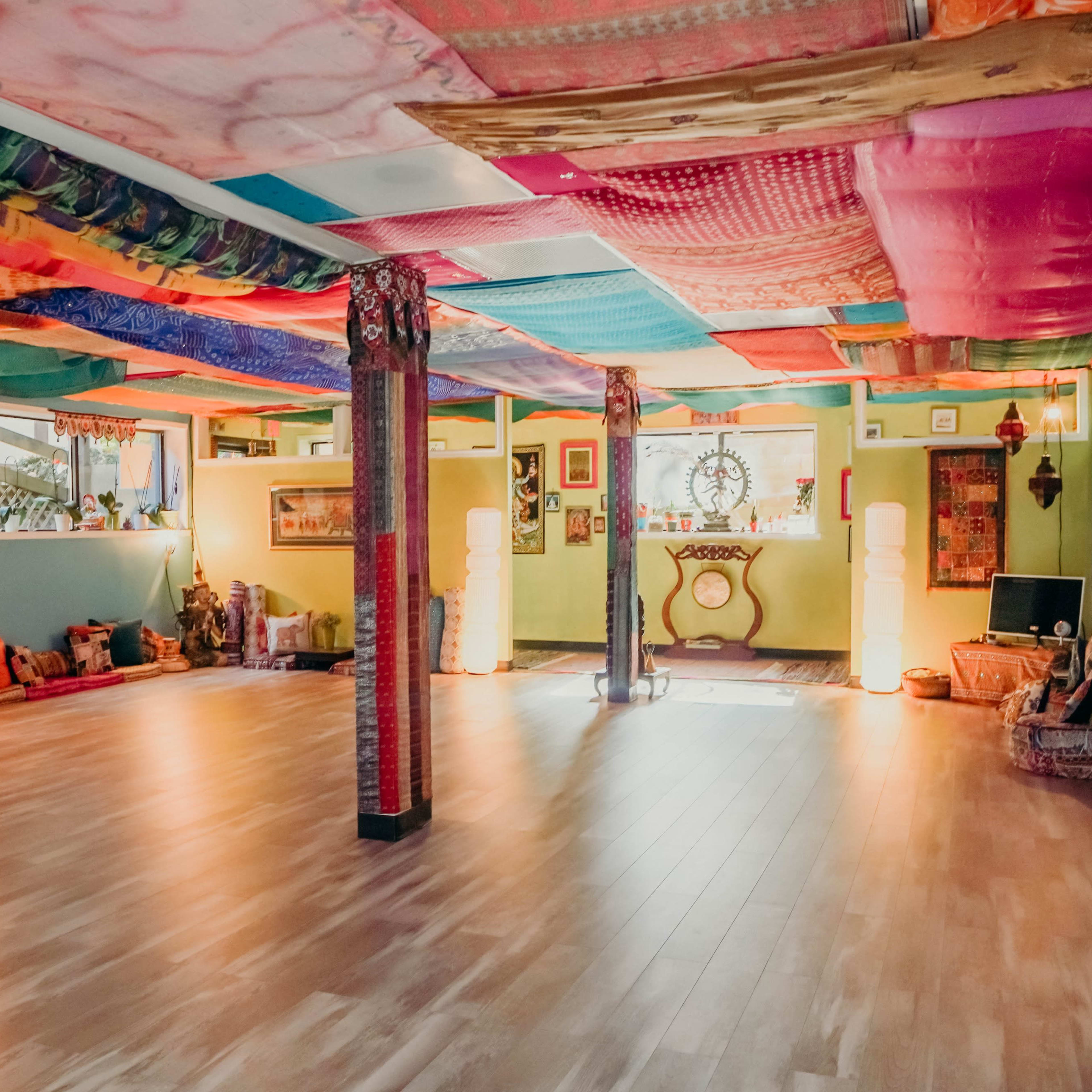 A brightly colored, spacious room with a wooden floor, adorned with vibrant fabric draping from the ceiling and illuminated by soft lamps.