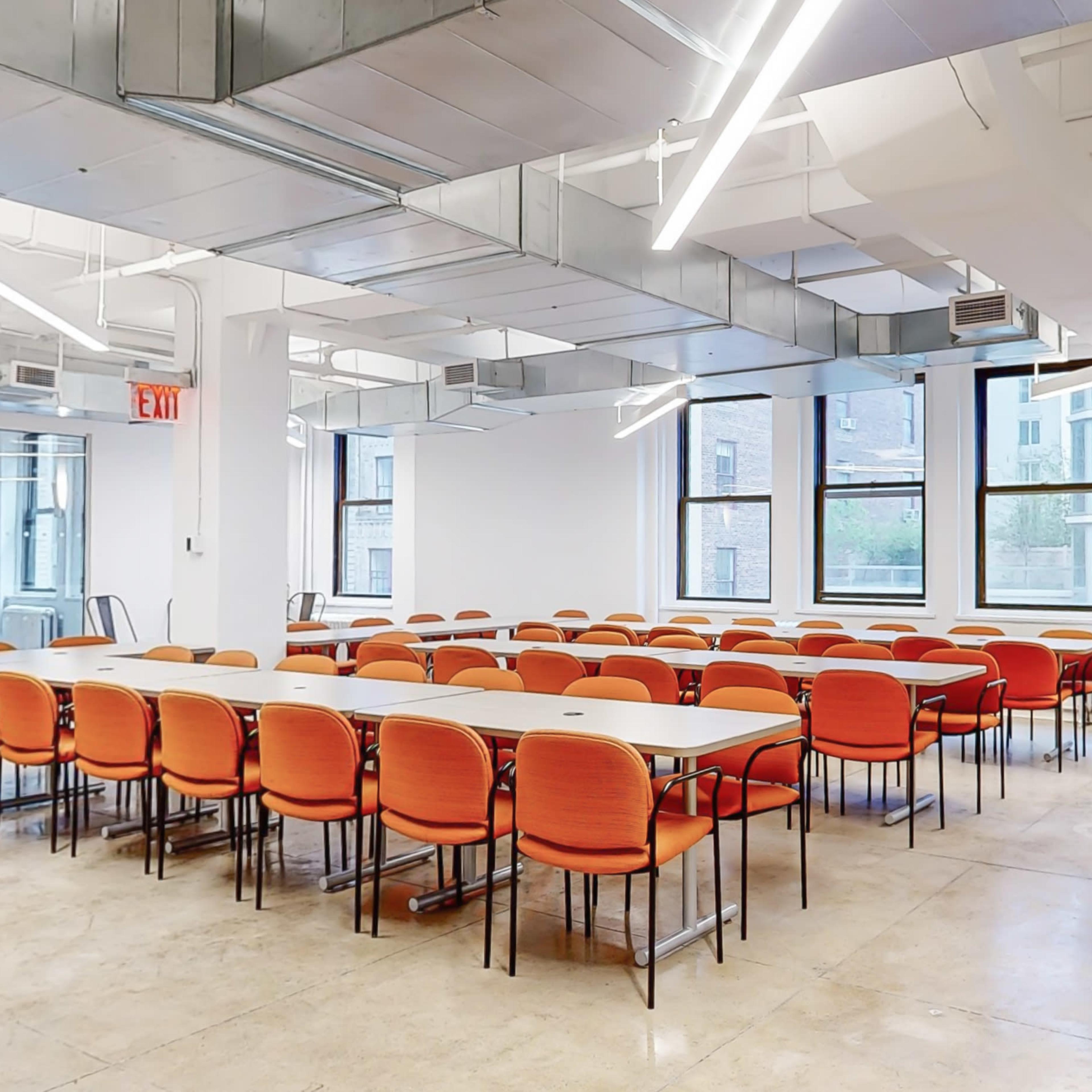 A modern conference room with a long table surrounded by orange chairs, large windows, and bright overhead lighting.