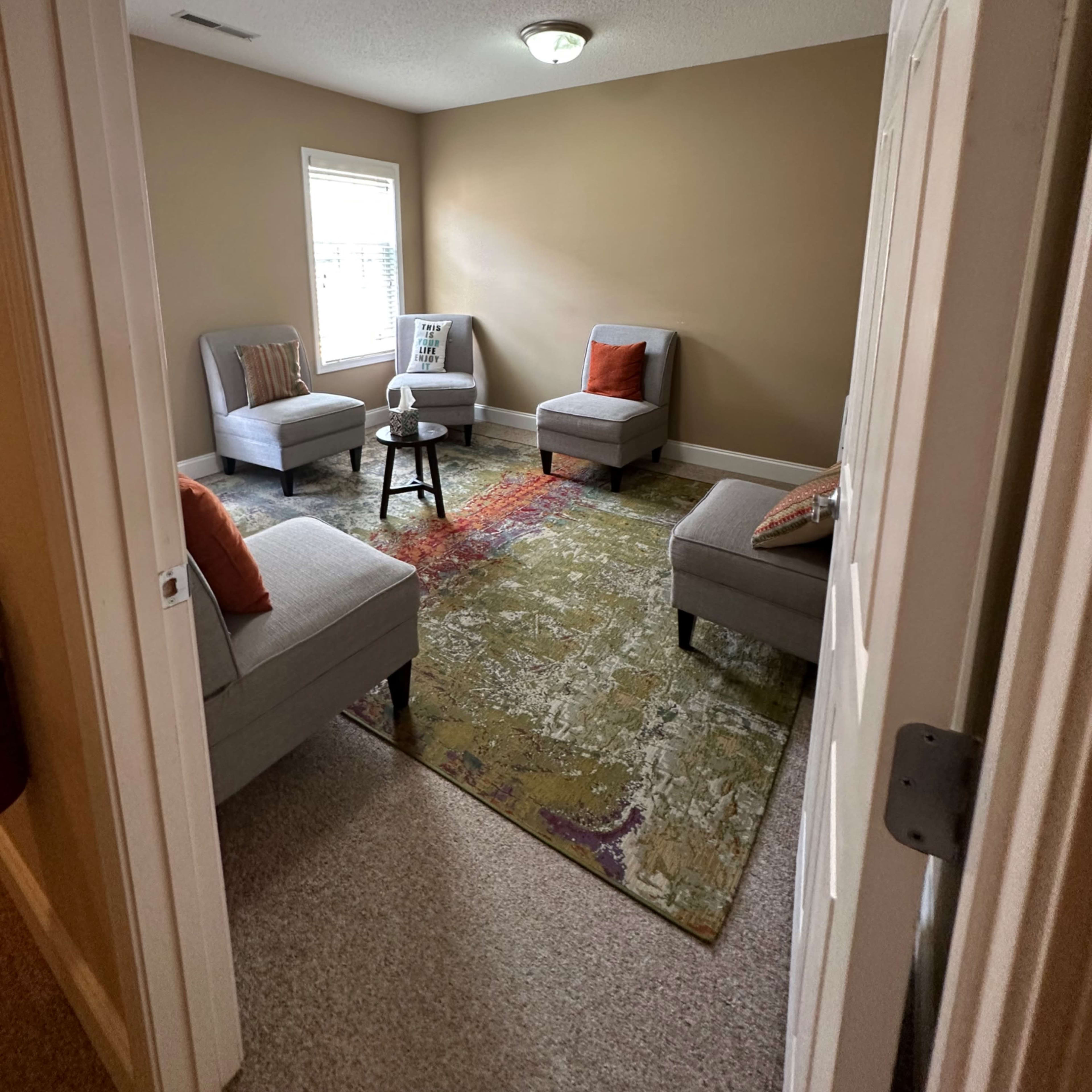 A small room with four gray chairs arranged around a small black table, with a colorful area rug on the floor and a window allowing natural light.