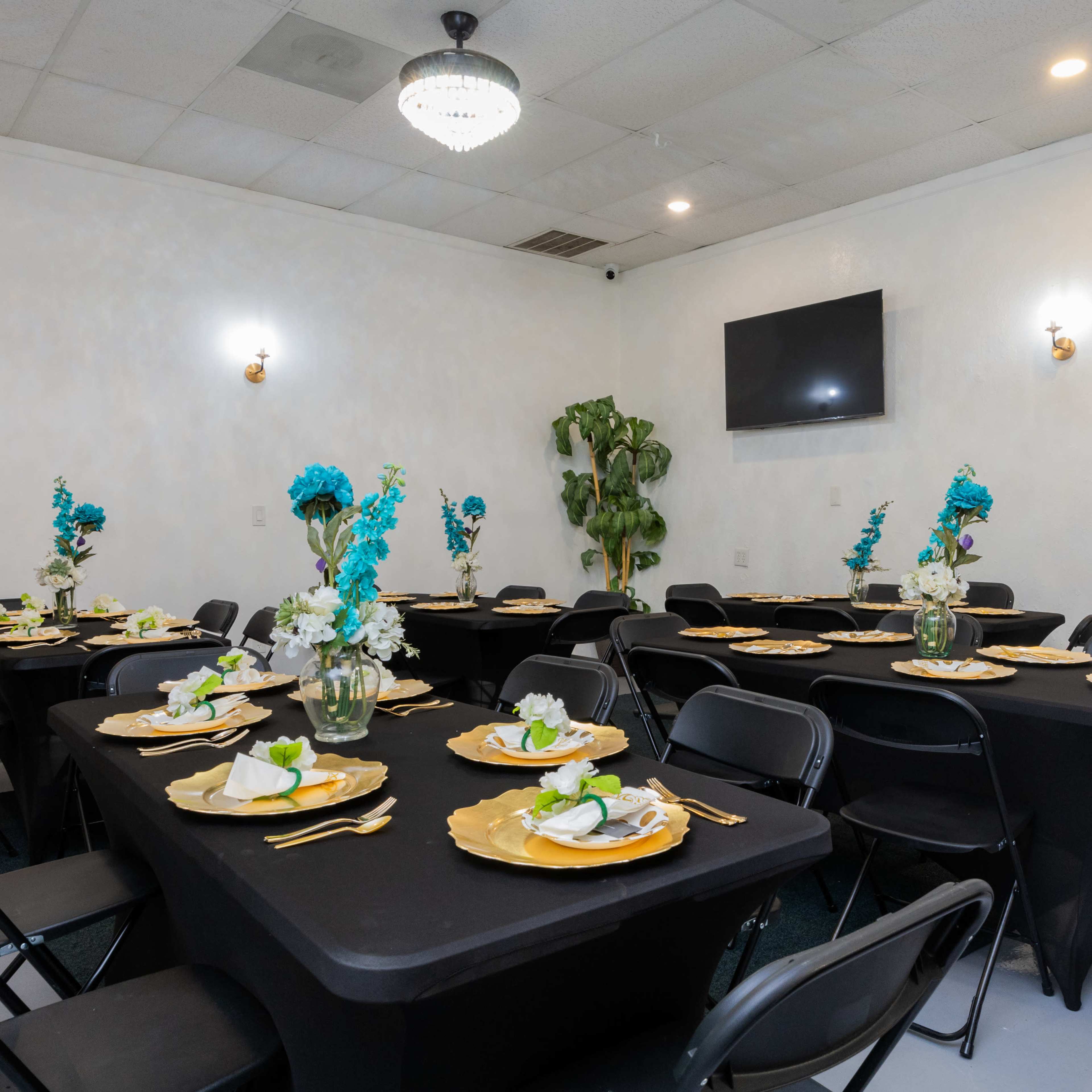 A dining room is set with black tables adorned with gold plates and white flowers, featuring two televisions mounted on the walls.