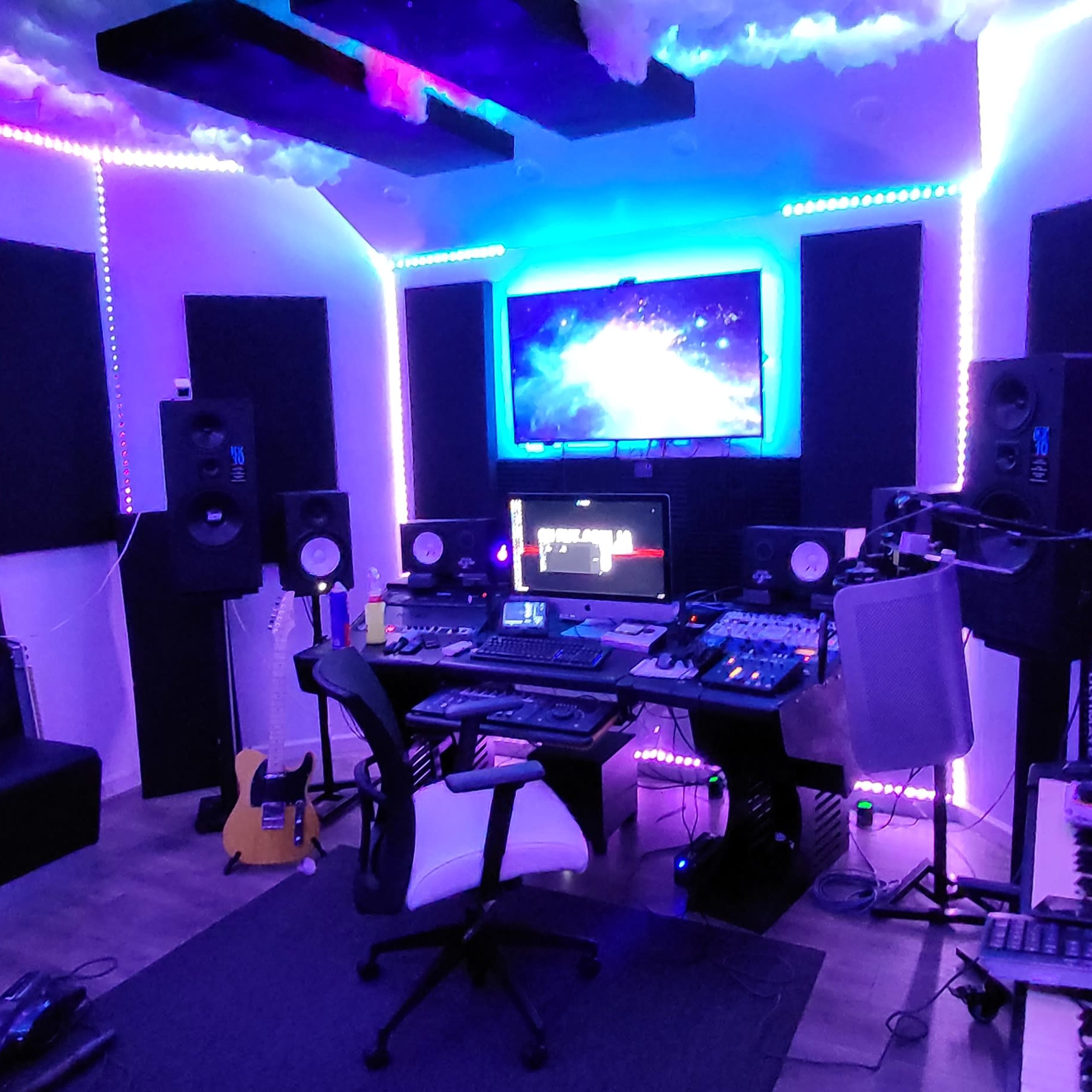 A music production studio features a central workstation with multiple screens, audio equipment, and various instruments, surrounded by sound-absorbing panels and LED lighting.
