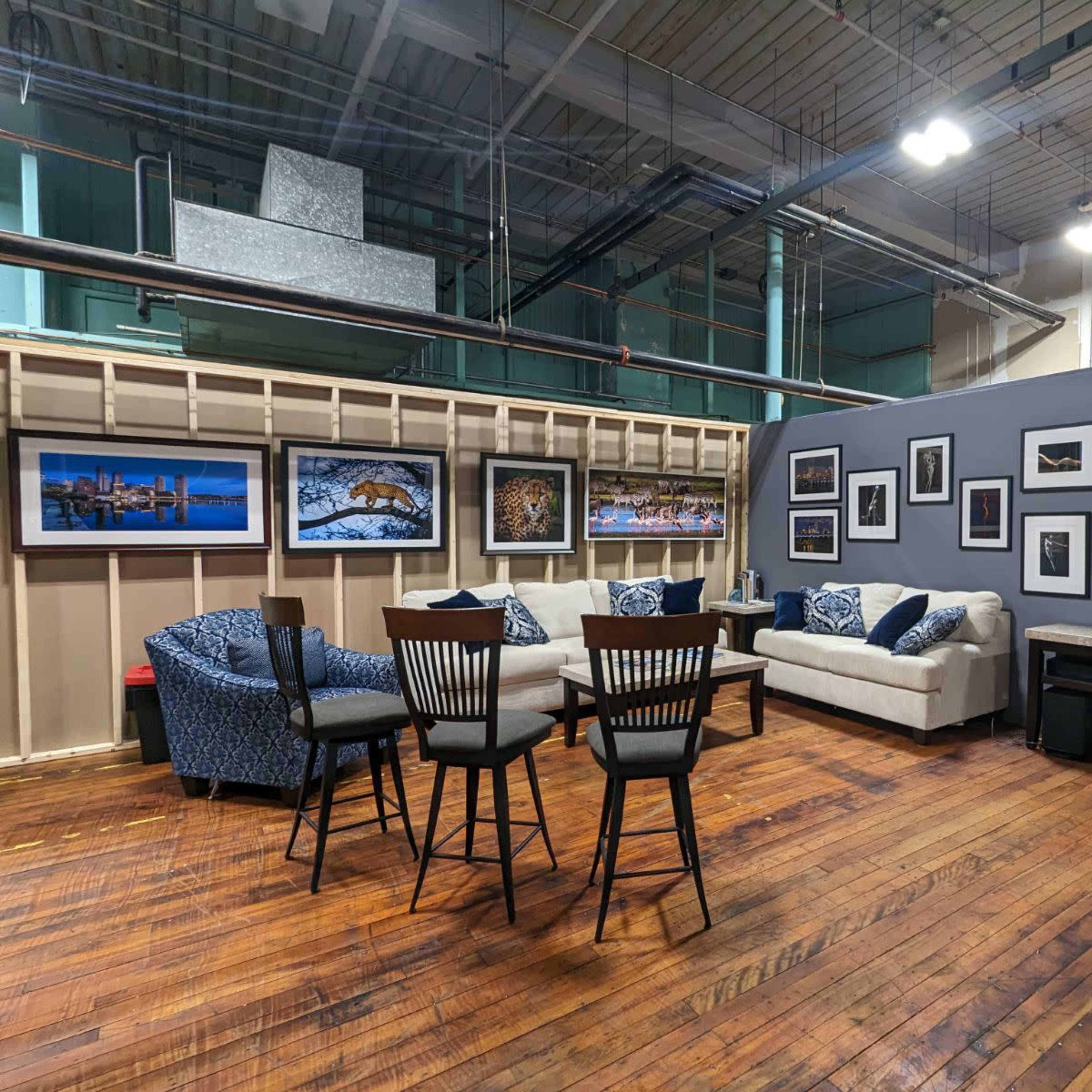 A modern seating area with a sofa and chairs arranged around a wooden table, surrounded by framed photographs on the walls in a spacious, industrial-style room.