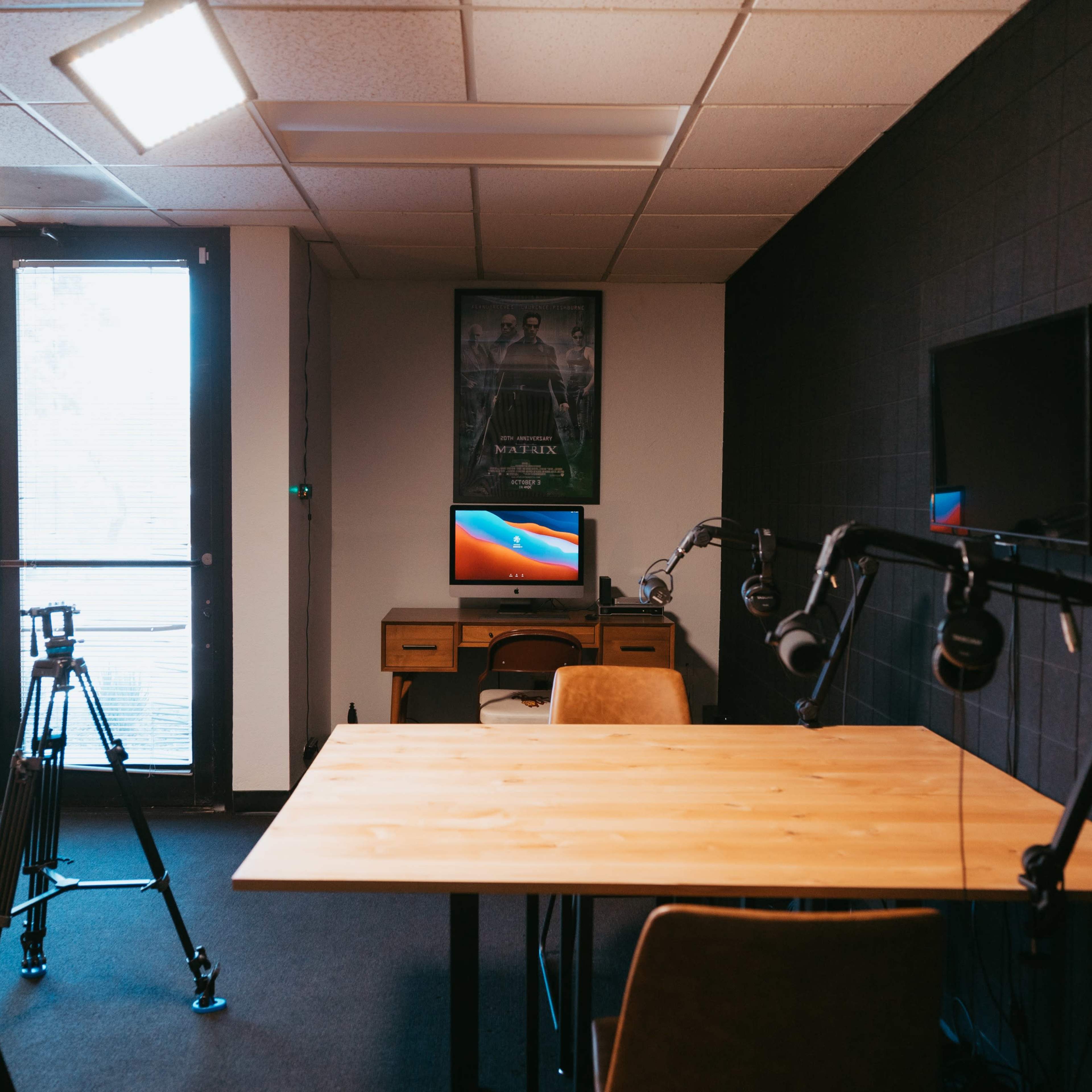 A modern recording studio with a wooden table, chairs, and camera tripods set up near a large window and a wall-mounted TV.