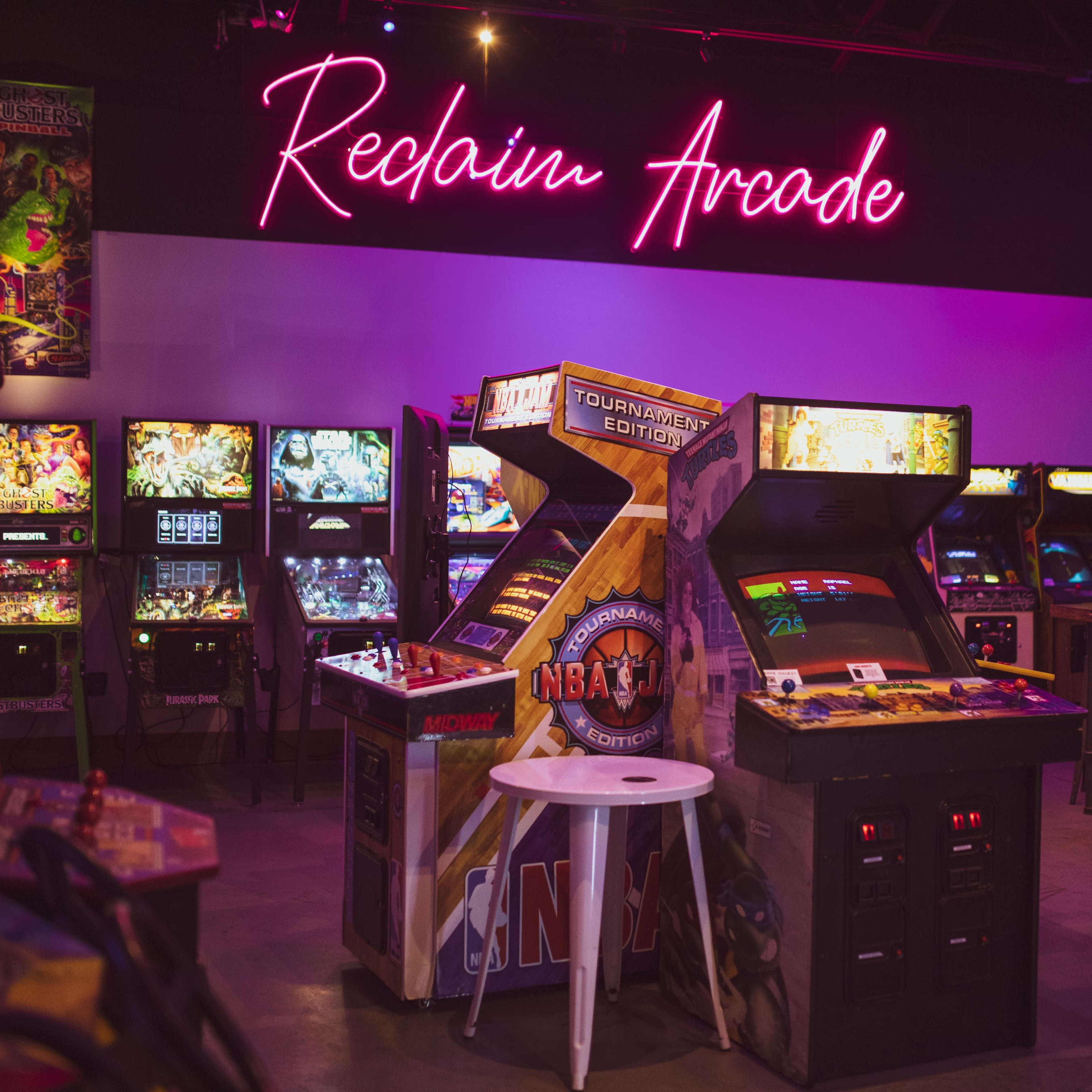 A vibrant arcade with several vintage gaming machines and a glowing neon sign that reads "Reclaim Arcade."